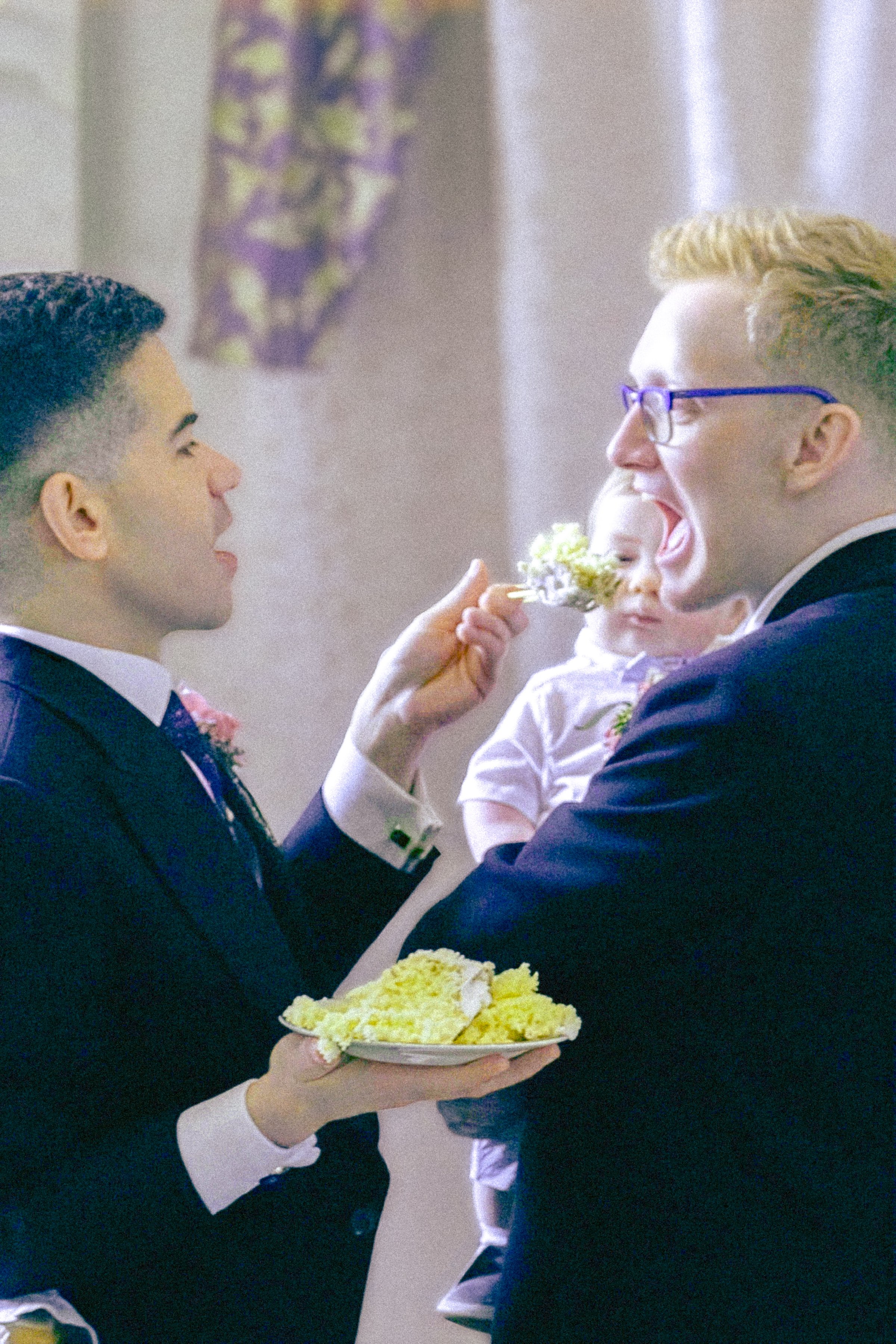 Two men in formal attire with boutonnières on their lapels, one holding a plate of yellow cake, and the other feeding a young girl a piece of cake, at a celebration or wedding event.