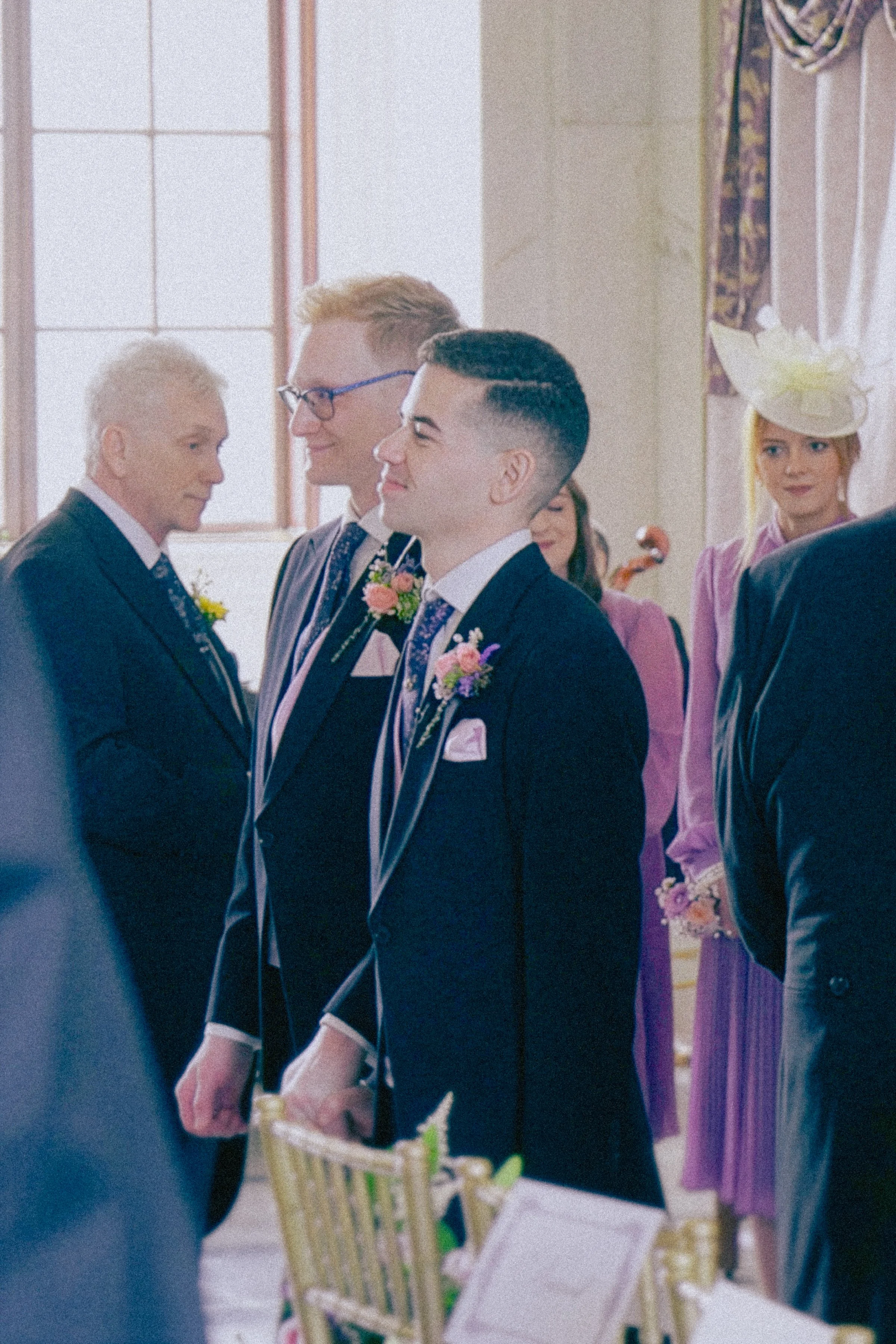 A wedding ceremony with groom and bride holding hands, surrounded by bridesmaids and groomsmen in formal attire inside a well-lit room with large windows.