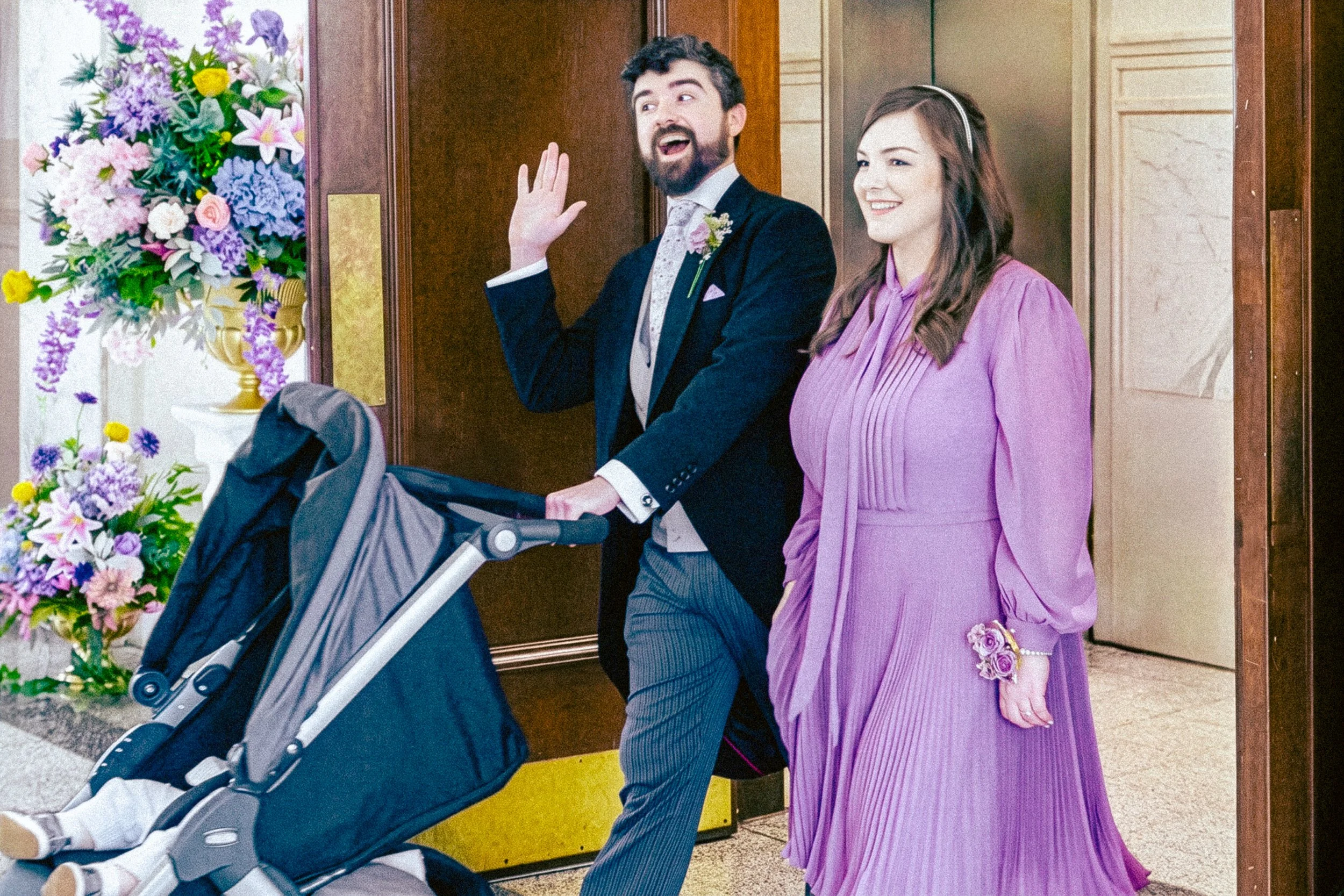 A man with a beard and a woman in a purple dress standing in an elevator lobby, smiling and waving while pushing a stroller, with a large bouquet of colorful flowers on the side.