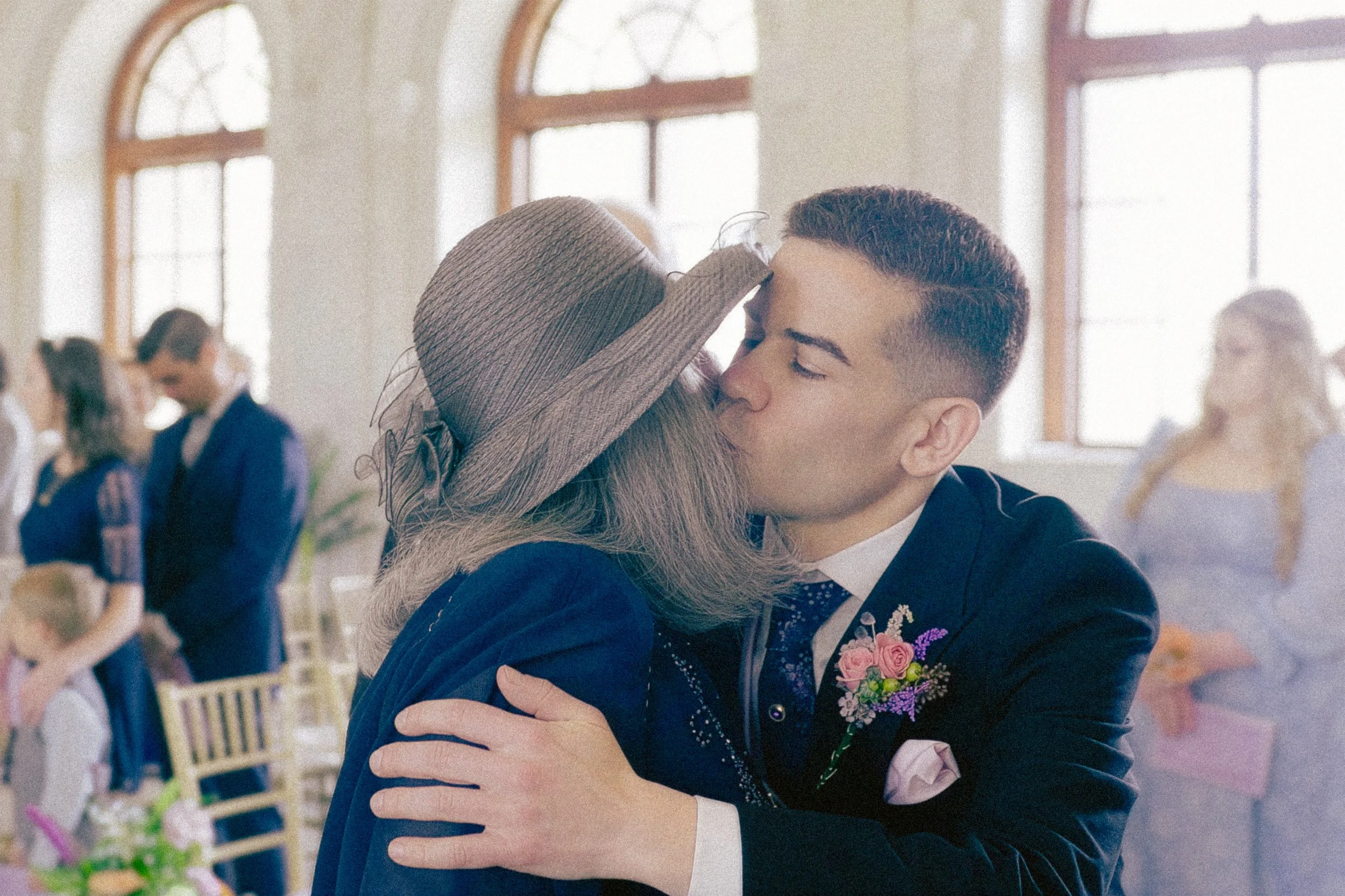 A young girl in a dress is hugging a man in a suit, who has a boutonniere on his lapel, inside a brightly lit room with large arched windows. The girl is wearing a large sunhat and the man has short dark hair. There are other people in the background
