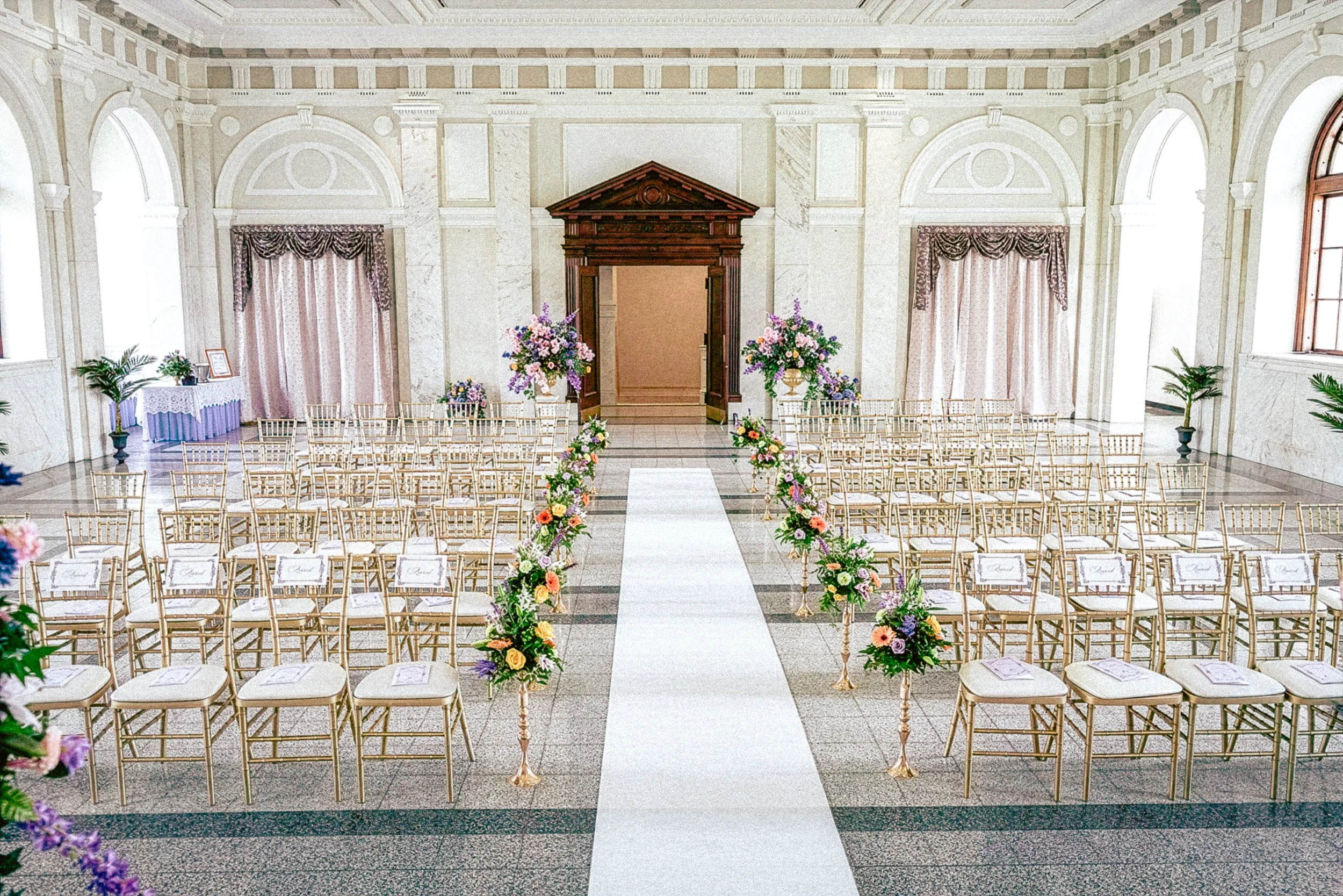 Wedding ceremony setup inside a grand hall with rows of gold chairs, floral arrangements, a white aisle runner, and a wooden arch at the front.