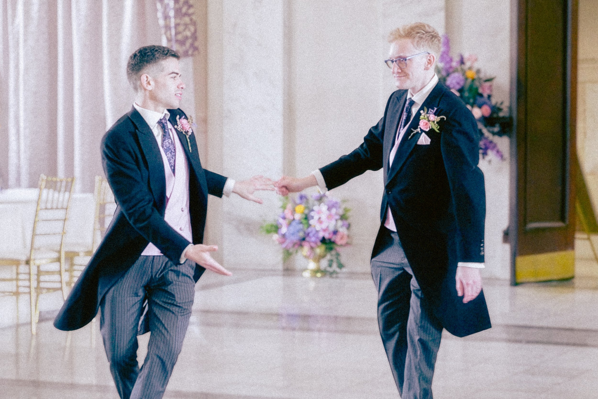 Two men in suits with boutonnières exchanging vows and holding hands at a wedding ceremony in a decorated indoor venue.