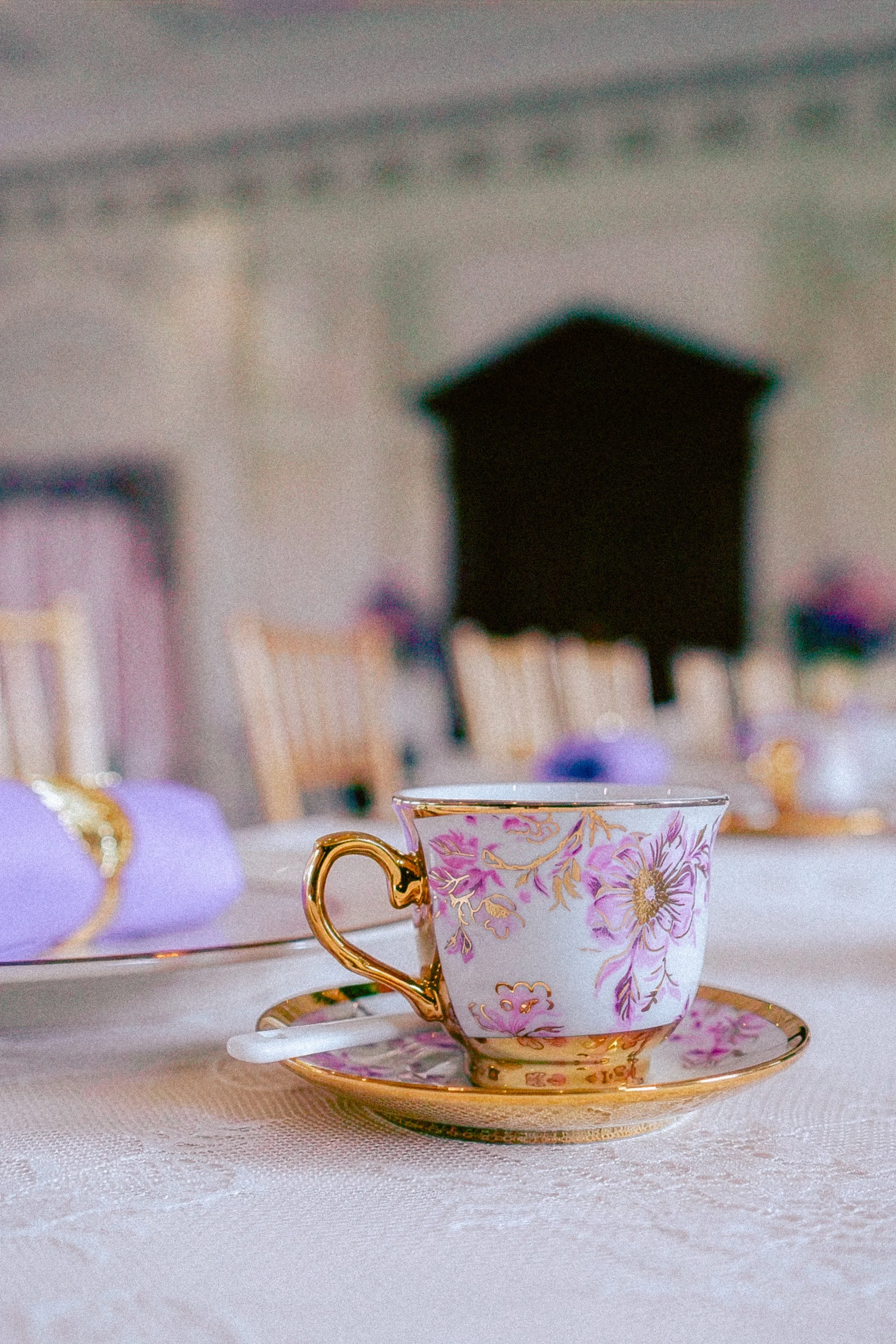 Elegant teacup with floral design and gold accents on a saucer, placed on a white tablecloth. In the blurred background, a dark shape possibly a speaker, and other colorful objects are visible.