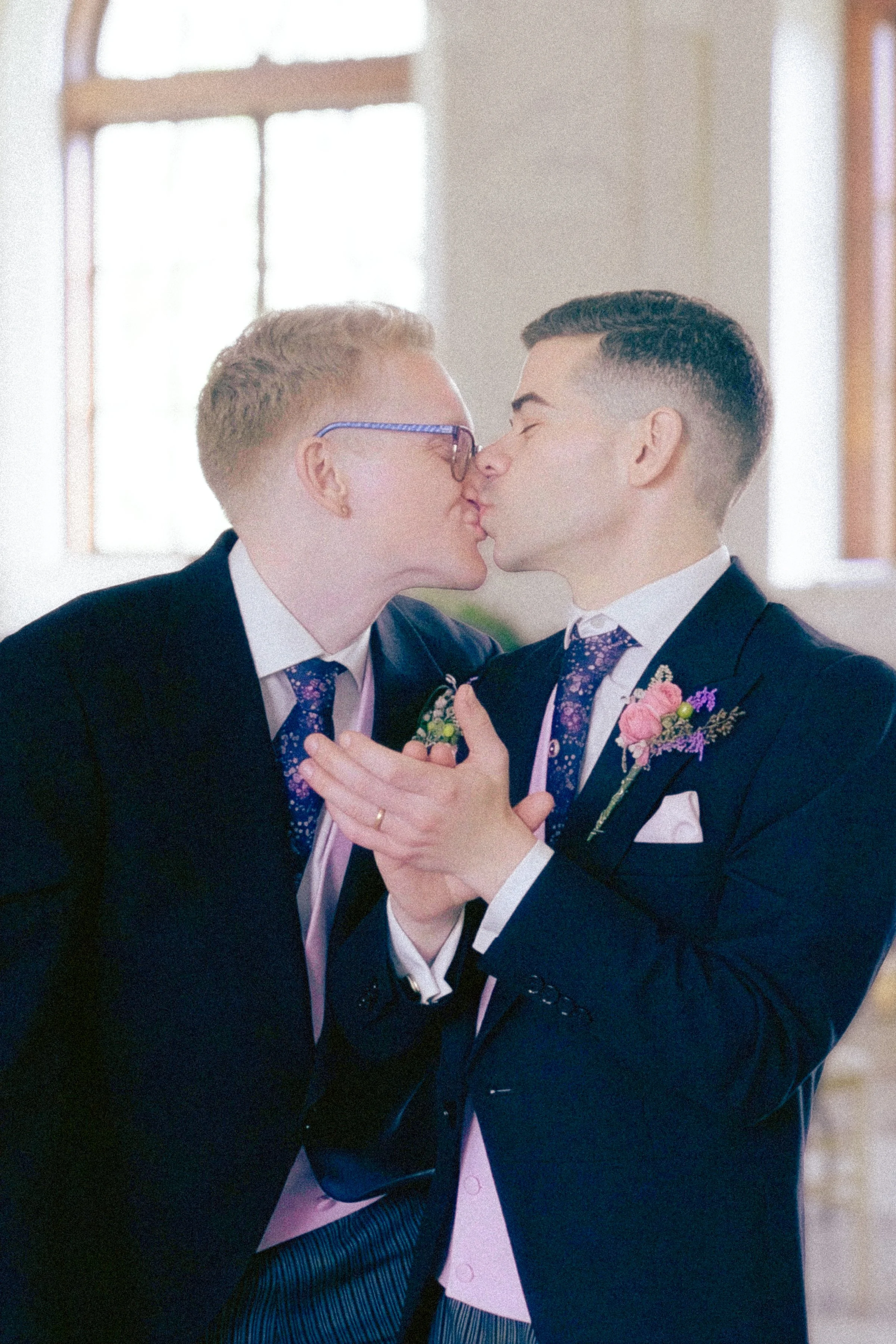 Two men in suits sharing a kiss at a wedding, holding hands, with a window in the background.