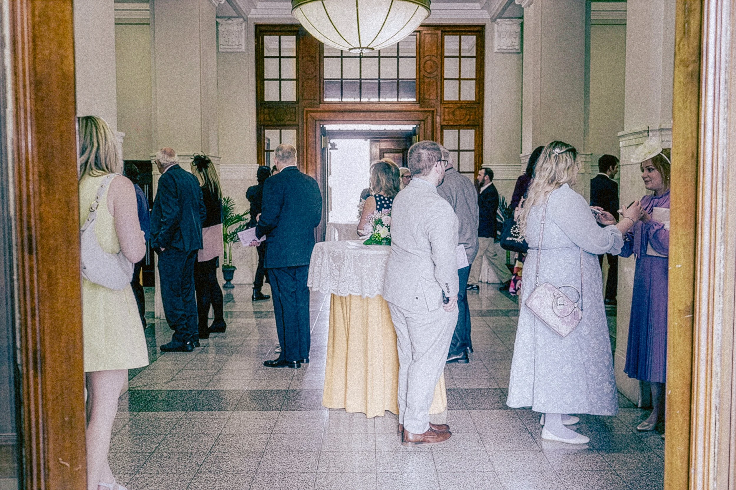 A group of people dressed in formal attire gathered in a hallway of an elegant building, possibly for a wedding or special event, seen through a large mirror.