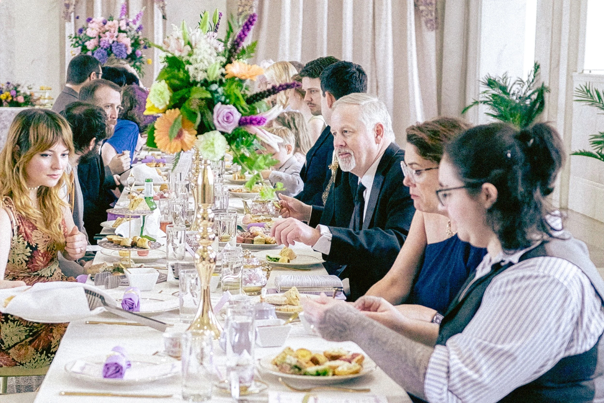 A long banquet table set for a formal event with a large floral centerpiece, surrounded by people eating and talking, in a decorated hall with curtains.