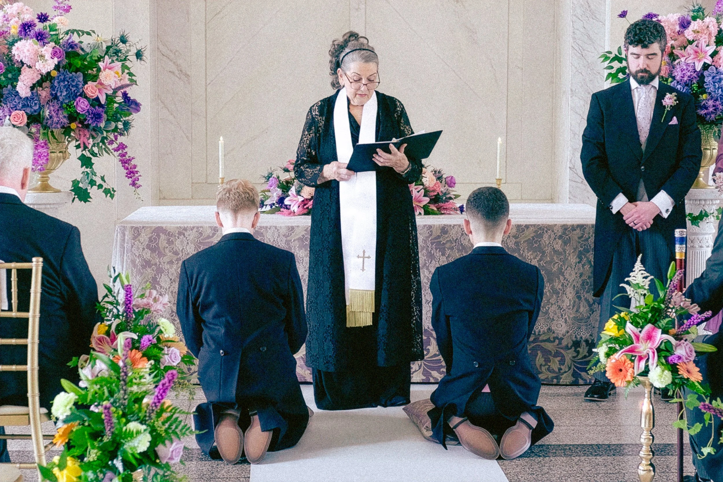 A wedding ceremony with pottoed couple kneeling, a officiant reading from a book, and three men in suits kneeling, surrounded by floral arrangements.