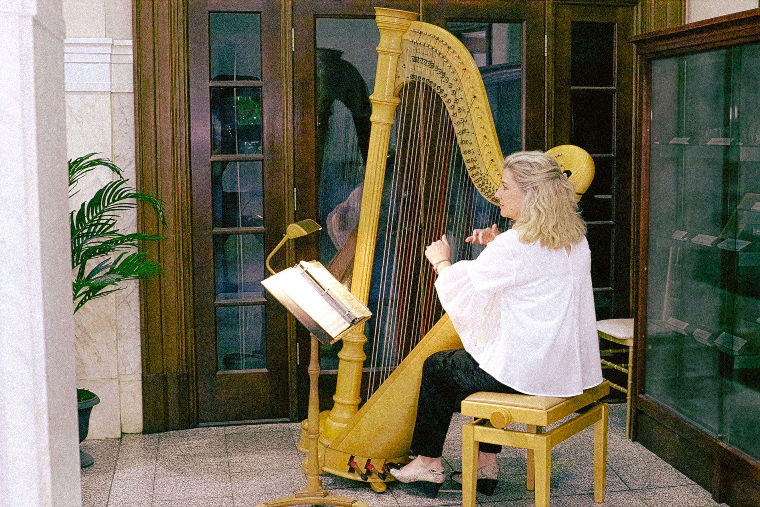 A woman playing a yellow harp indoors, seated on a yellow bench, with a music stand in front of her and a glass display case on her left.