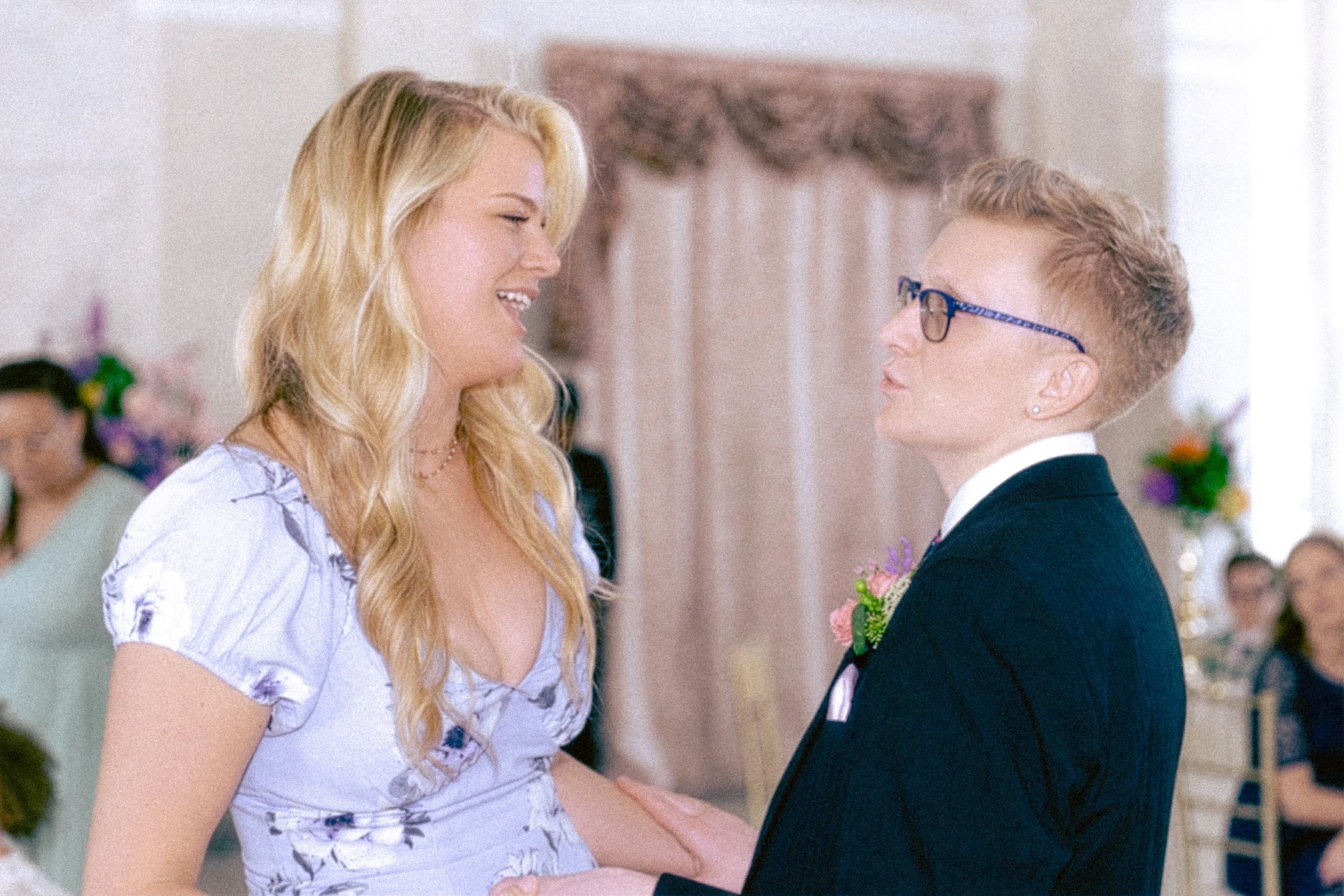 A young woman and a young man are holding hands and looking at each other, possibly during a wedding ceremony in an indoor setting with people and flowers in the background.