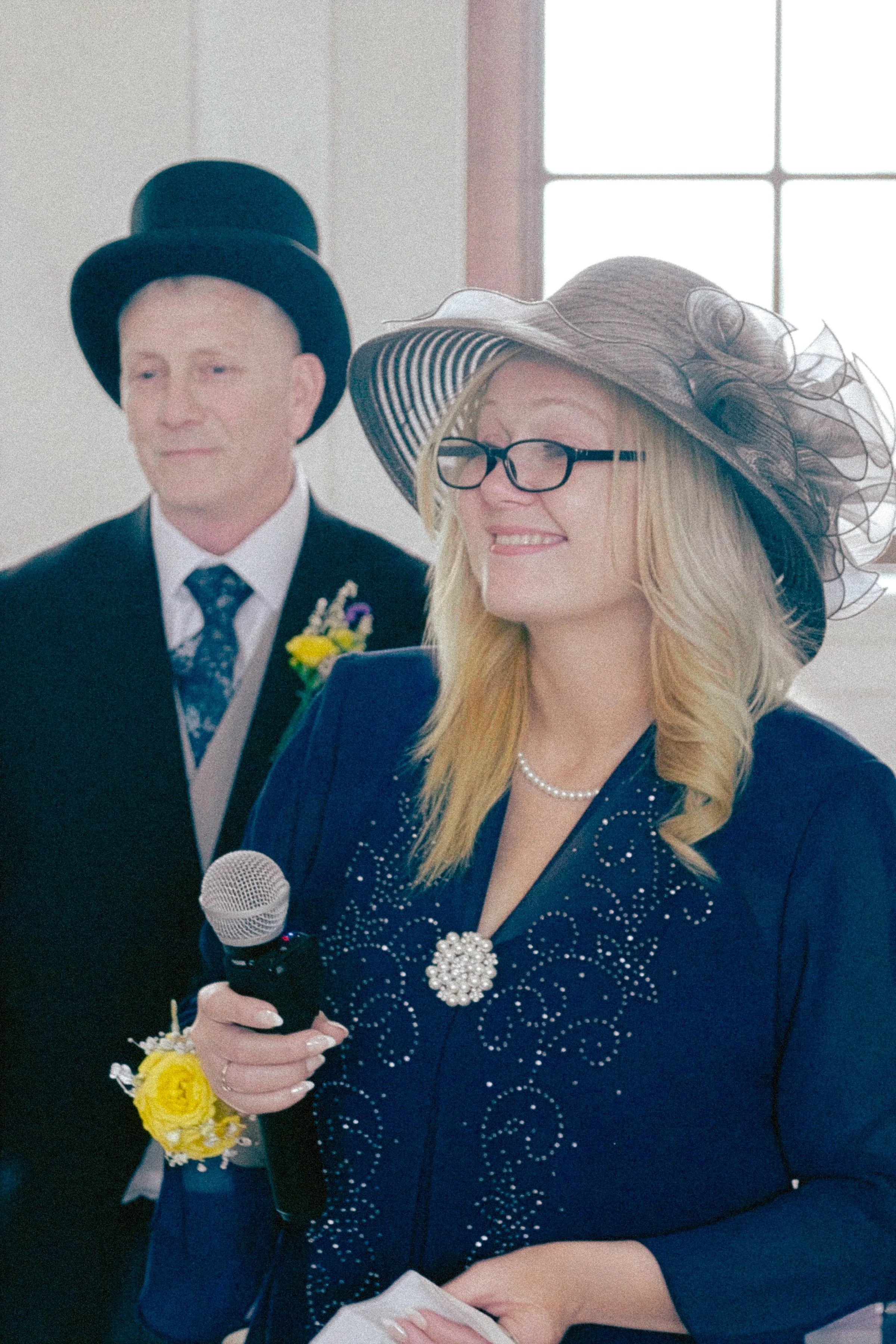 A woman dressed in blue with pearl jewelry, wearing a large black and beige hat with decorative netting, is smiling and holding a microphone during an event. A man in a suit, tie, and top hat, with a boutonniere, stands behind her next to a window.