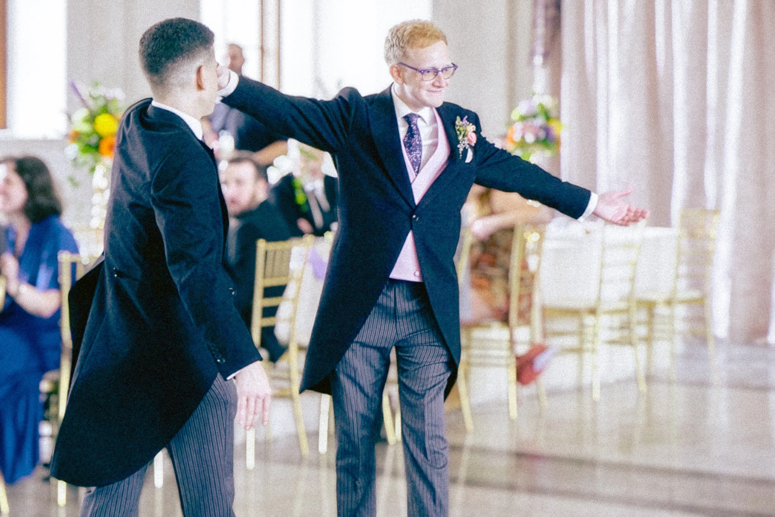 Two men in formal attire dancing at a wedding reception, with floral arrangements and guests seated in the background.