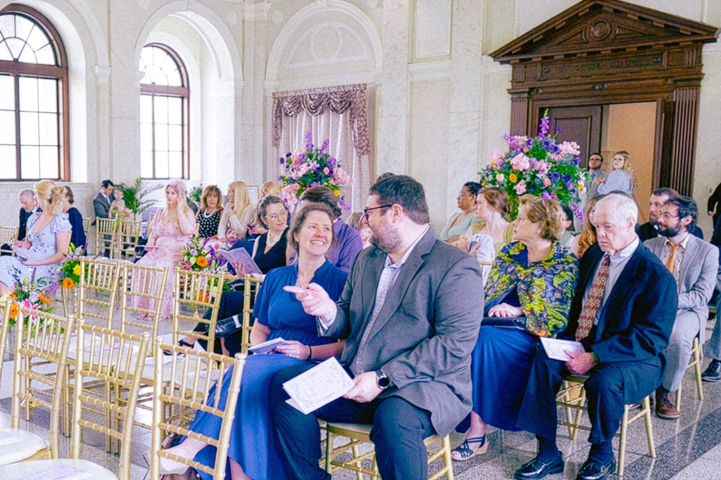 People attending a wedding ceremony seated in a decorated venue with floral arrangements and large windows.
