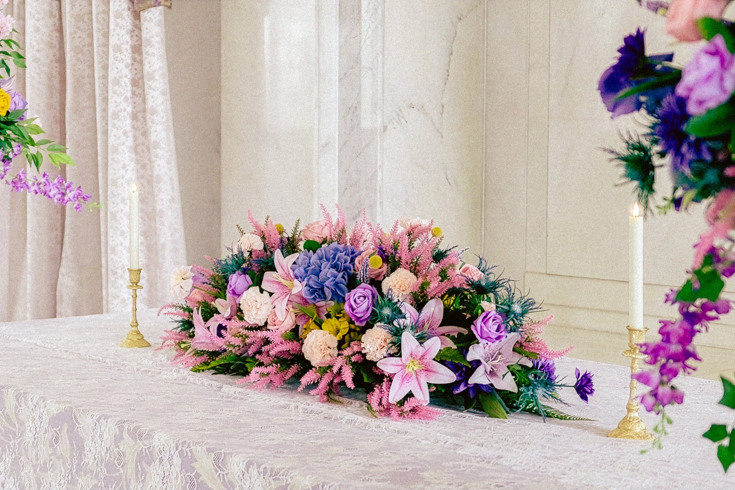 A floral centerpiece with pink, purple, and blue flowers on a white lace tablecloth, surrounded by gold candleholders with white candles, on a decorated table in a room with white walls and lace curtains.