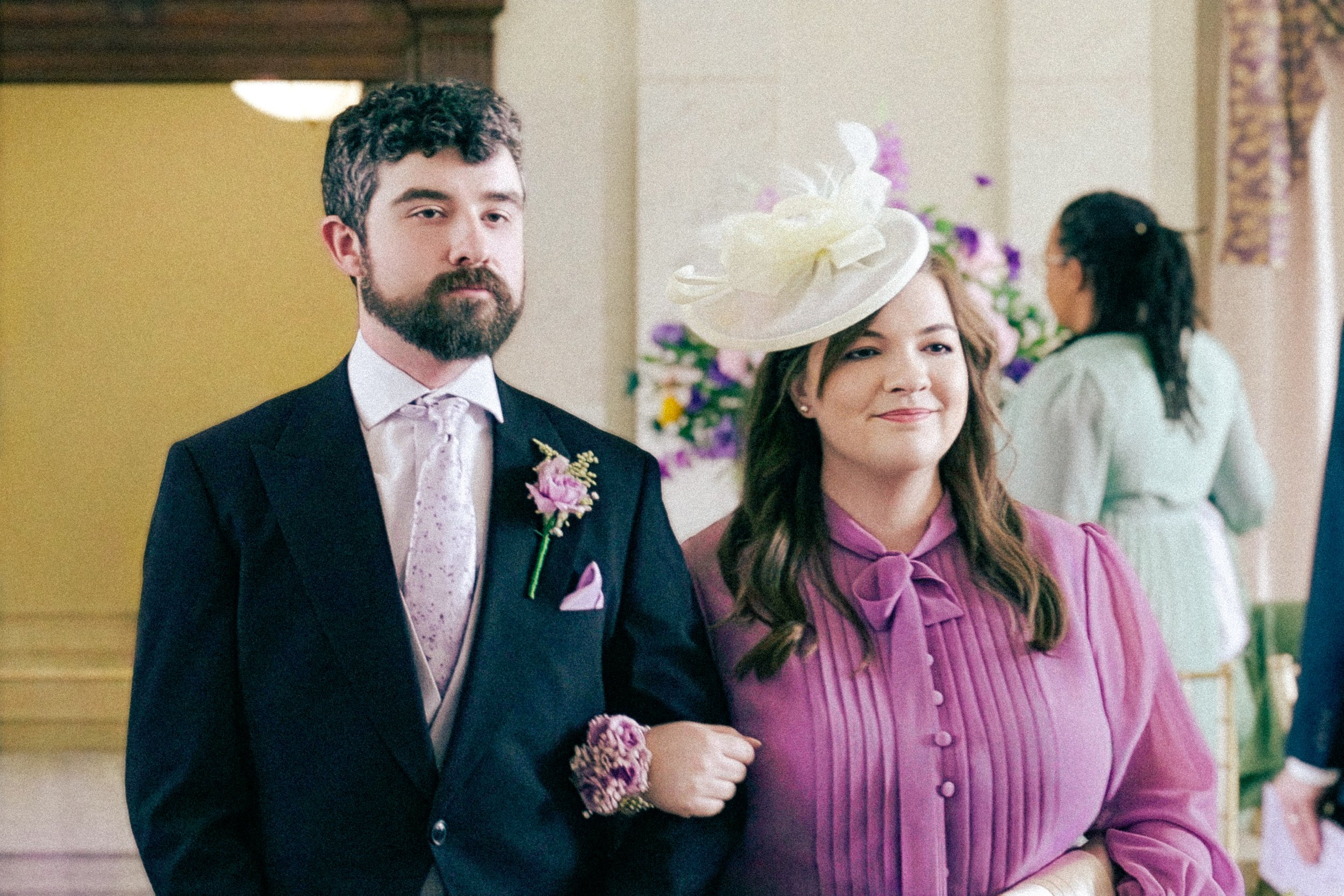 A man and woman dressed in formal attire, standing close together at an event, with the man in a black suit and tie and the woman in a purple dress with a large white hat. They are surrounded by others in the background and bulletin boards.