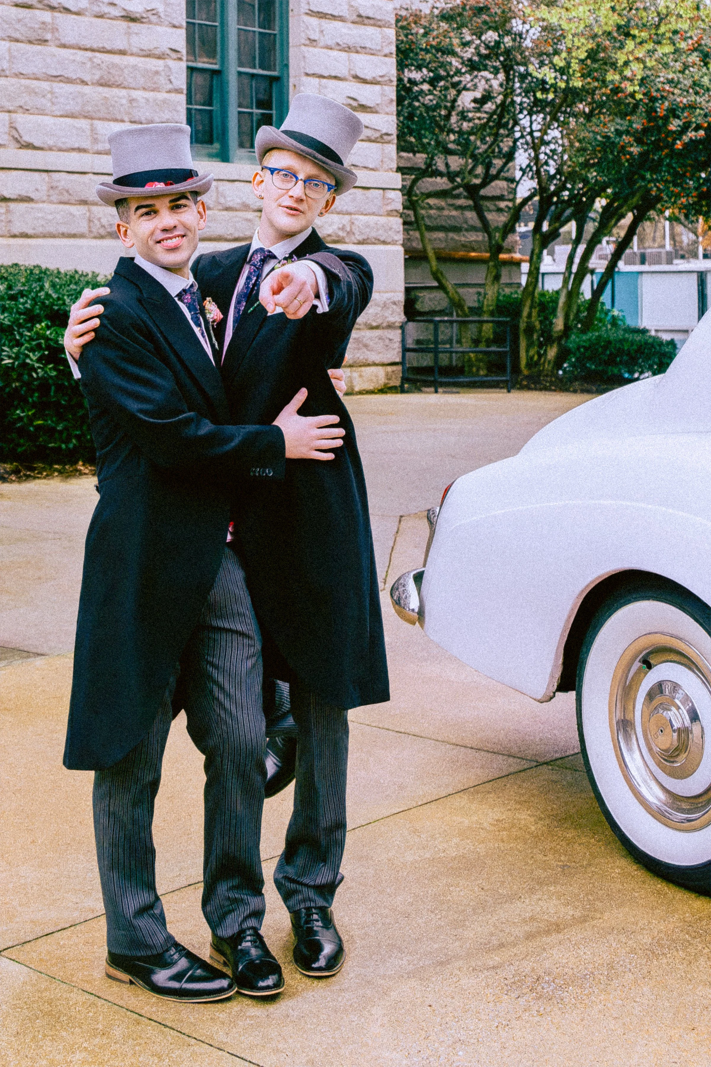 Two men dressed in tuxedos and top hats, standing close and smiling, with one man pointing toward the camera. They are outdoors next to a white classic car, on a paved surface with bushes and a stone building in the background.
