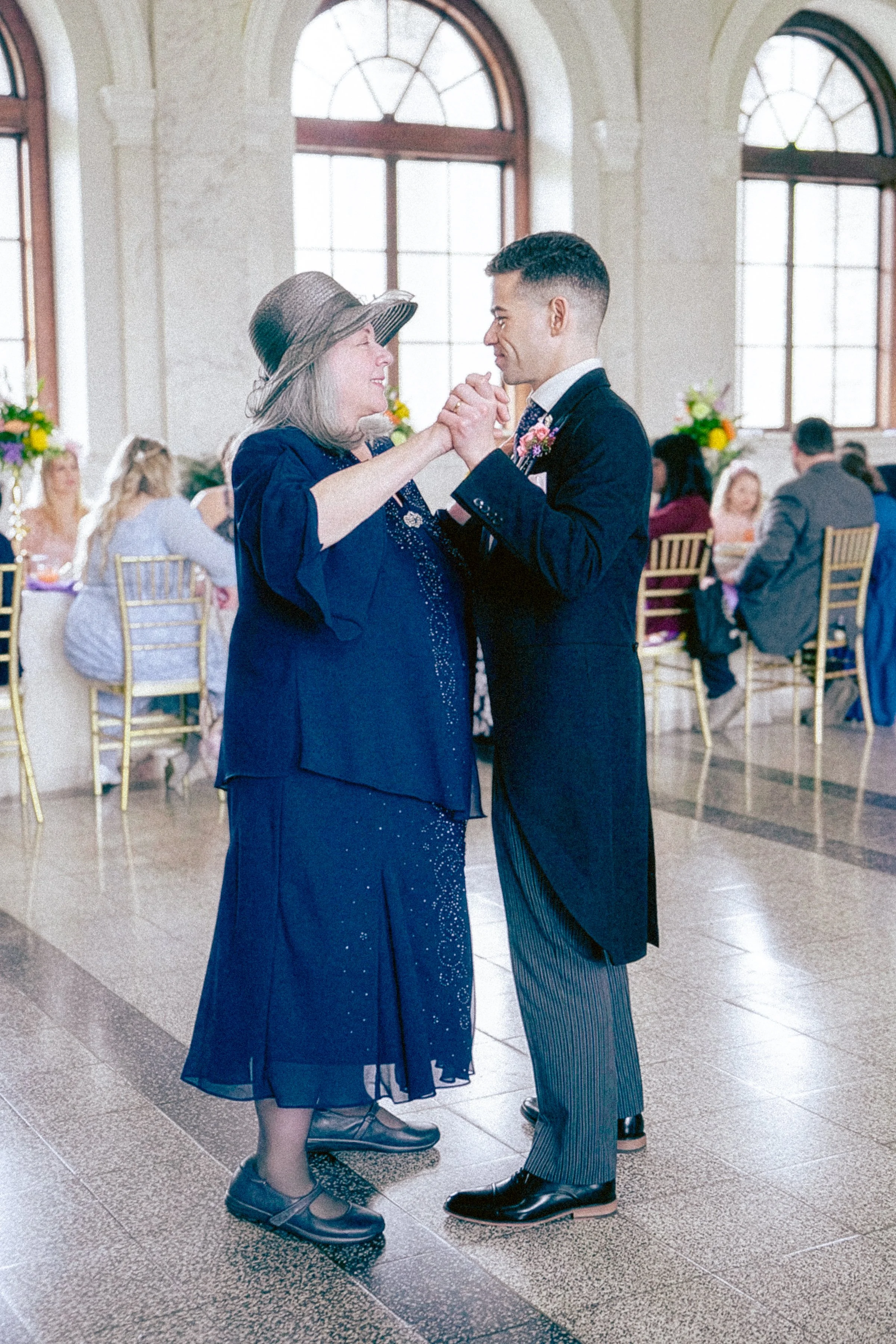 A woman and a man are dancing together in a bright, elegant hall with large arched windows. The woman is wearing a dark blue dress and a wide-brimmed hat, and the man is dressed in a black suit with pinstripe pants. They are holding hands and smiling
