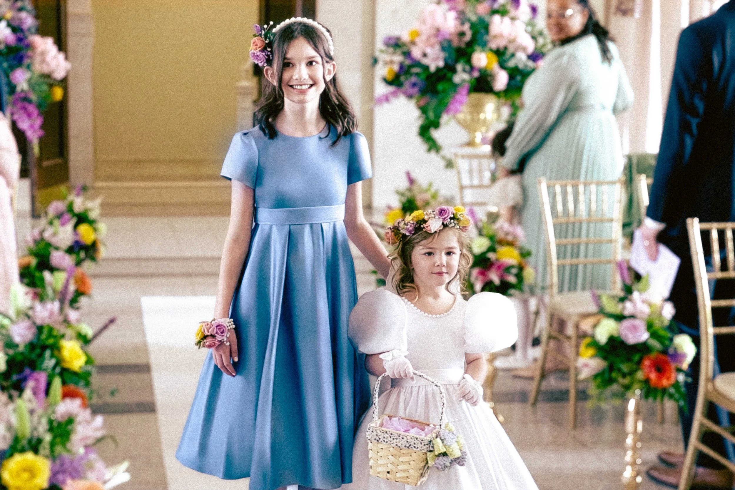 Two young girls, one in a blue dress and the other in a white dress, holding hands in a flower decoration setting, with floral arrangements in the background.