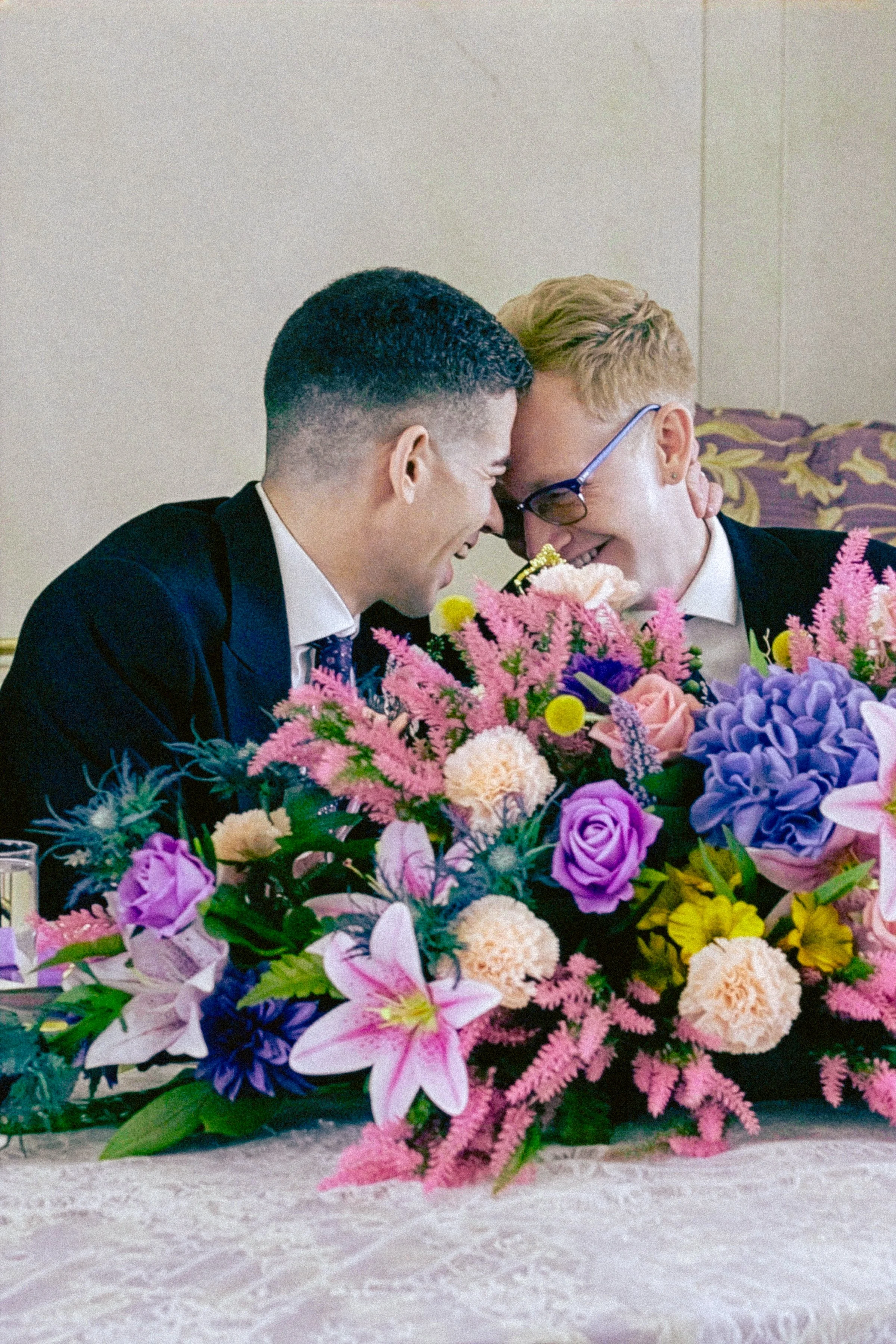 A vibrant bouquet of pink, purple, and peach flowers, including lilies, roses, and carnations, arranged on a table with a blurred background.
