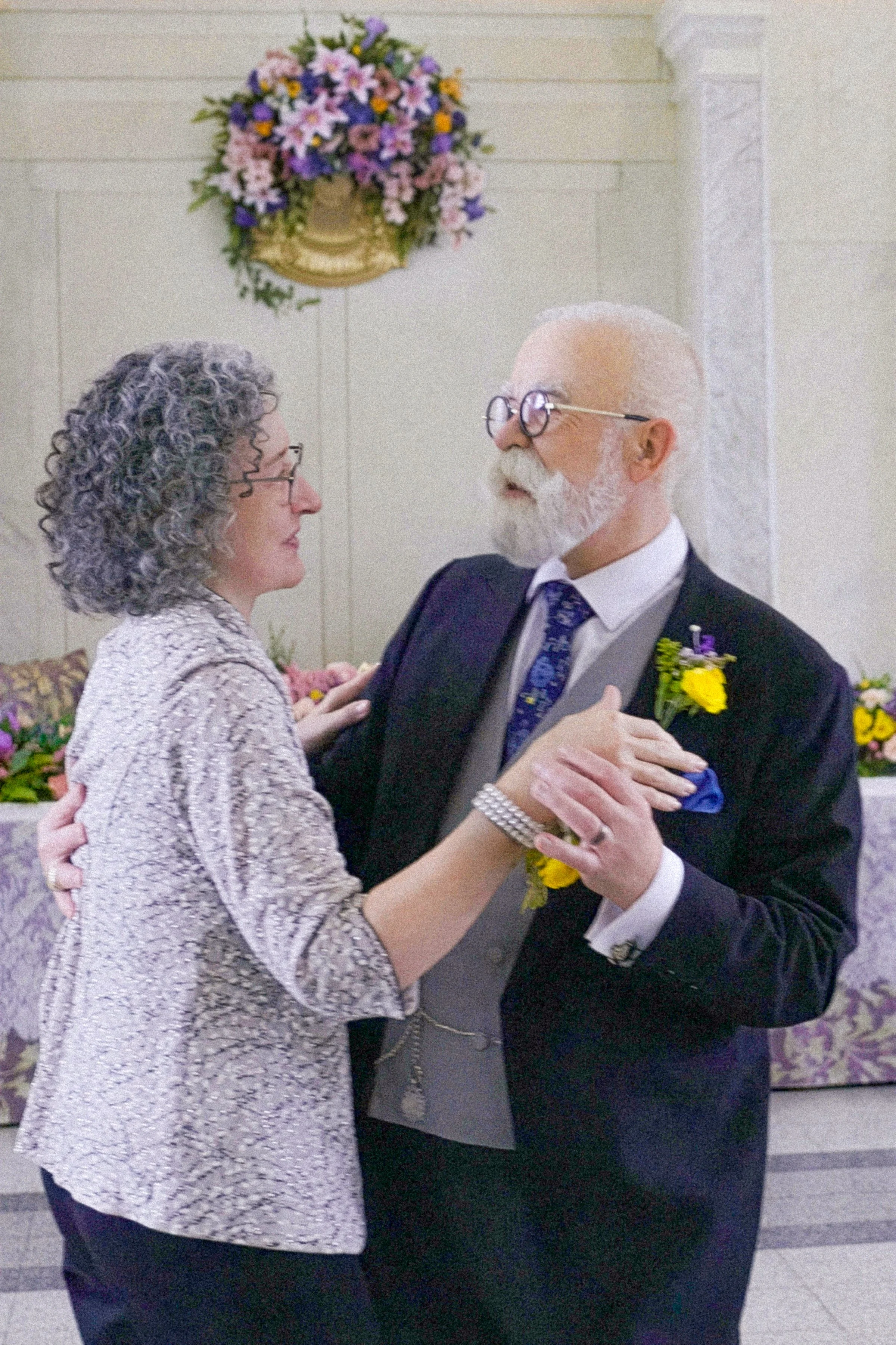 An elderly couple dancing at a wedding reception, with floral arrangements in the background.
