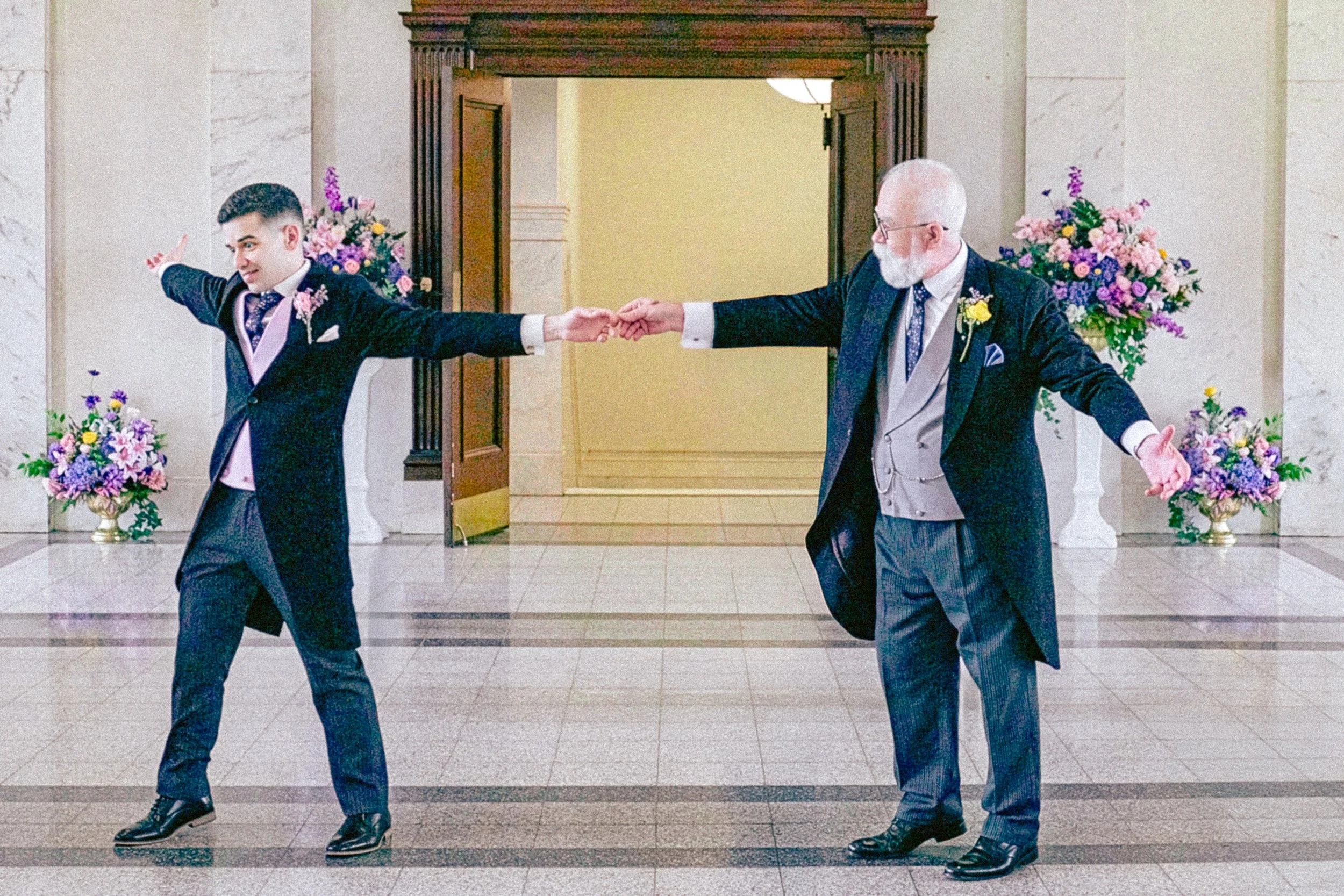 A young man and an older man are holding hands and dancing in a formal setting with floral arrangements.