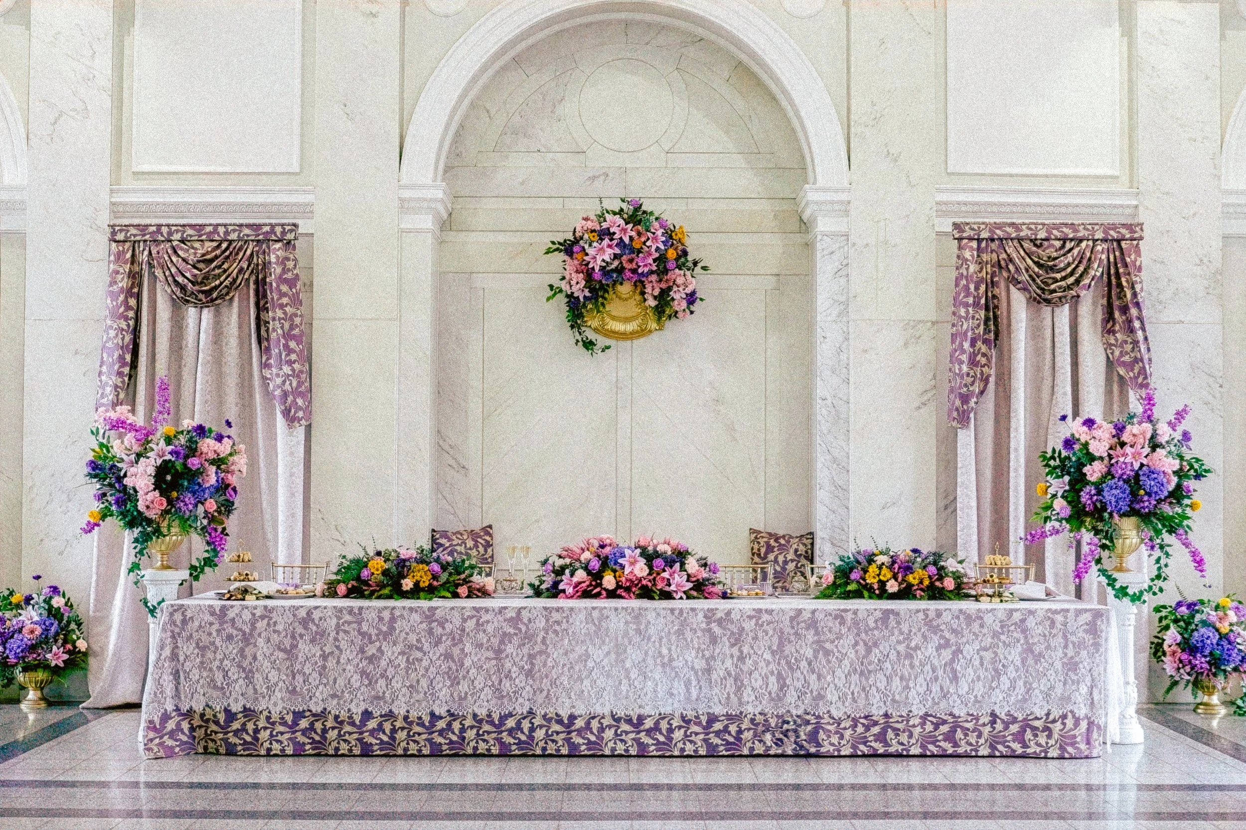 A decorated wedding or event reception table with purple and pink flowers, gold accents, and elegant draped curtains in a marble-walled hall.