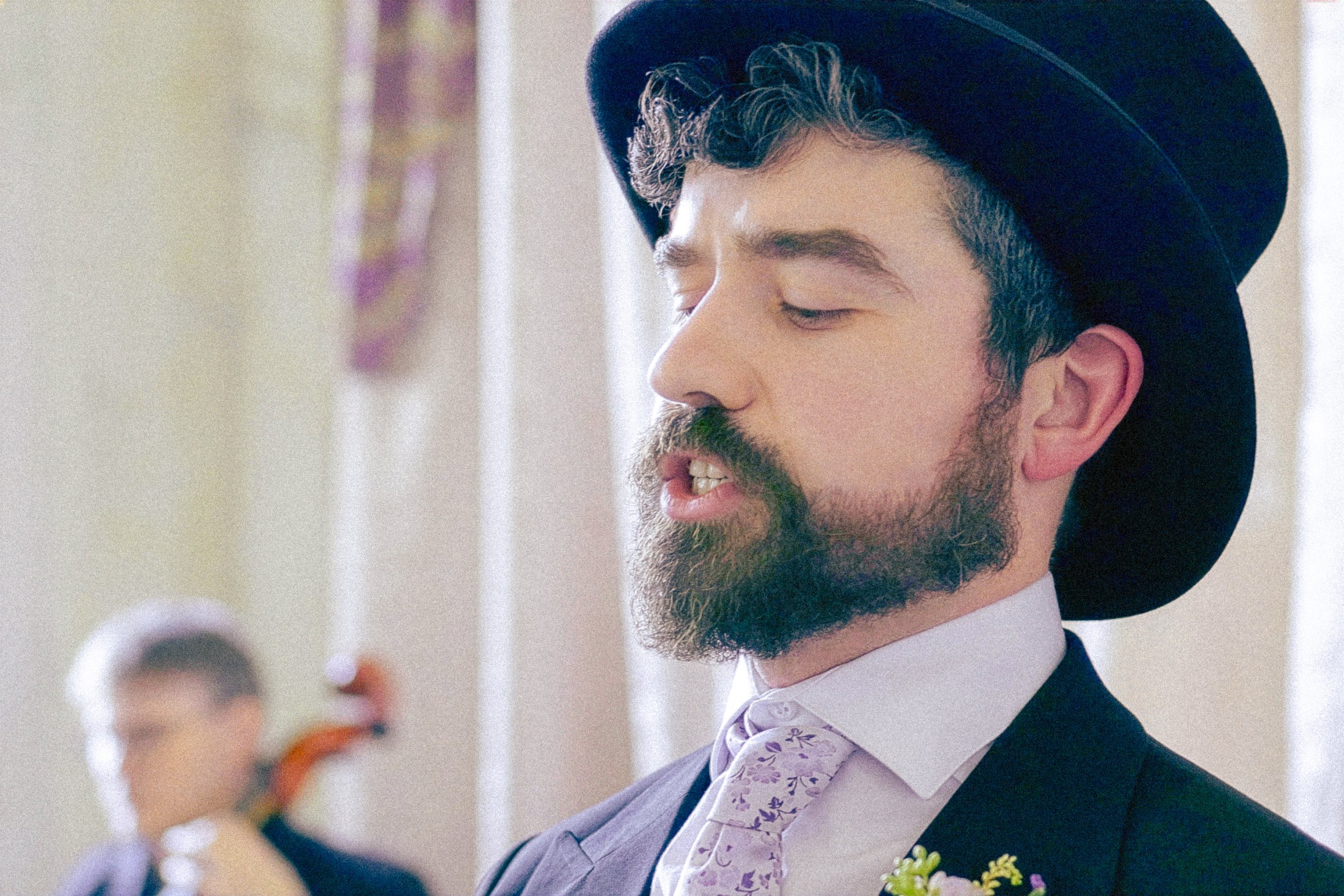 A man with a beard and mustache wearing a black hat, white shirt, purple patterned vest, and black suit jacket, looking downward with his eyes closed.