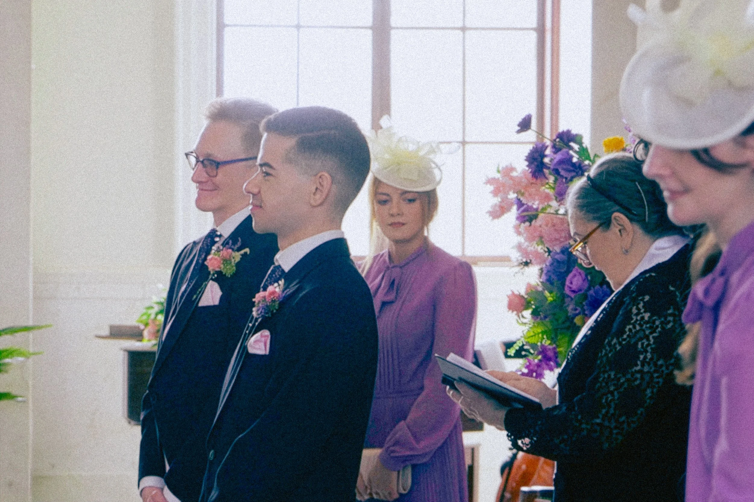 People attending a wedding ceremony, standing in line in a bright room with large windows, holding papers or programs, with floral arrangements in the background.