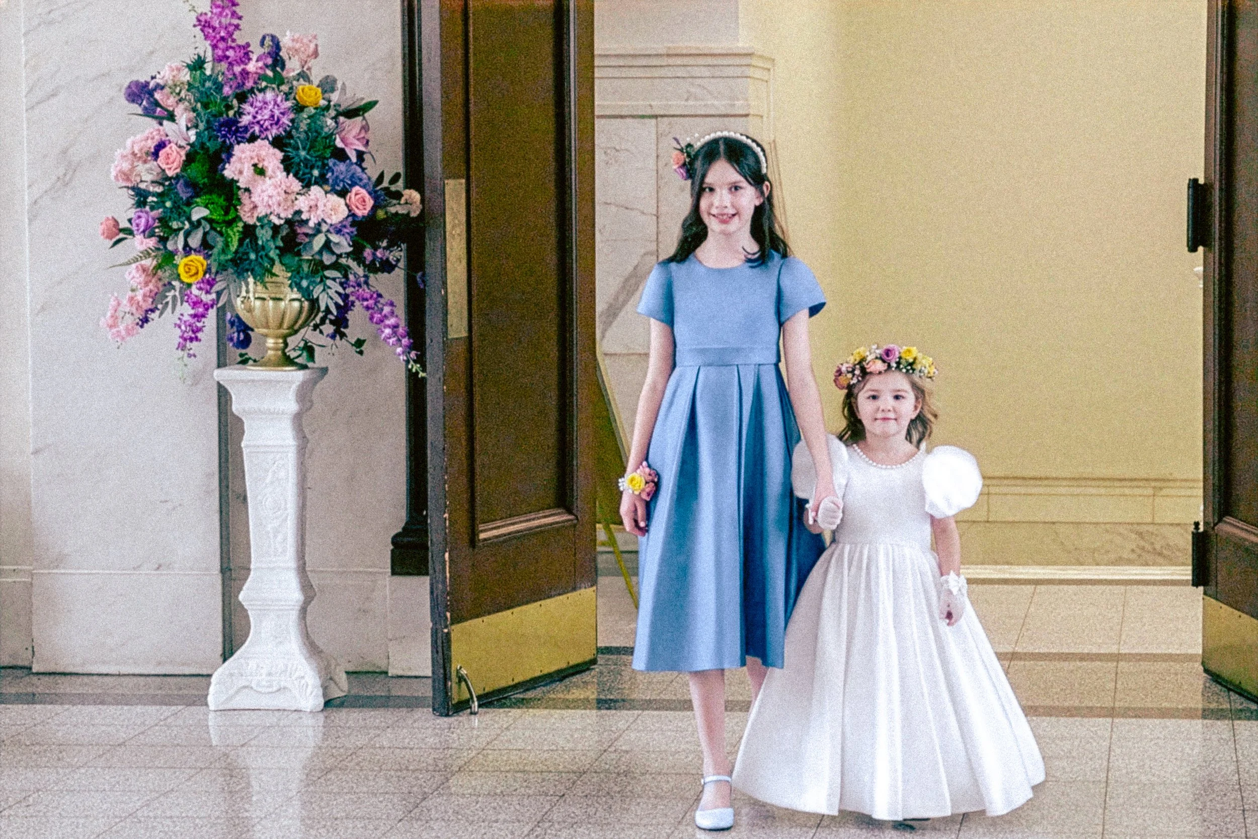 Two young girls dressed in formal attire, holding hands, and standing in front of an open door with a large floral arrangement on a white pedestal nearby.