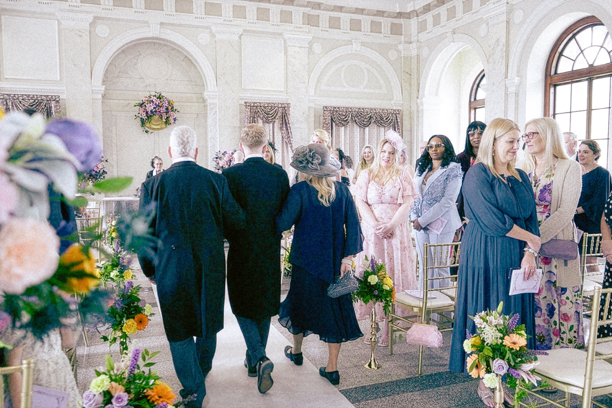 People attending a formal event in a grand, decorated hall with arched windows, floral arrangements, and gold chairs, dressed in elegant and semi-formal attire.