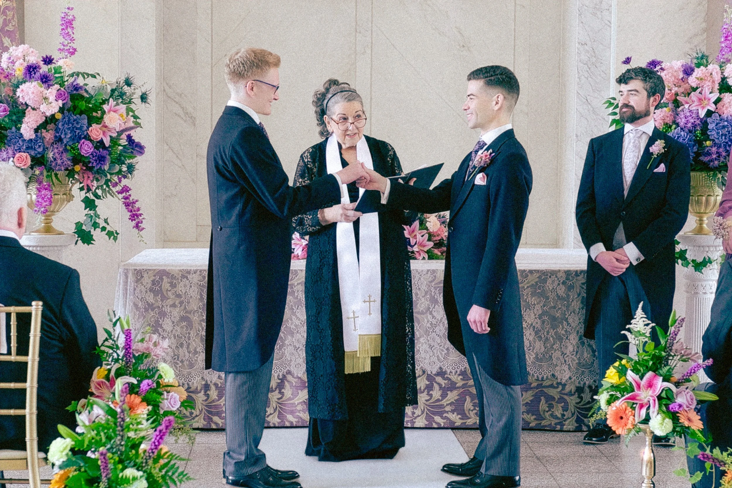 A wedding ceremony is taking place with an officiant and two grooms exchanging vows. The setting is decorated with large floral arrangements in pink, purple, and green, and the grooms are dressed in matching dark suits with pink flowers on their lapels.