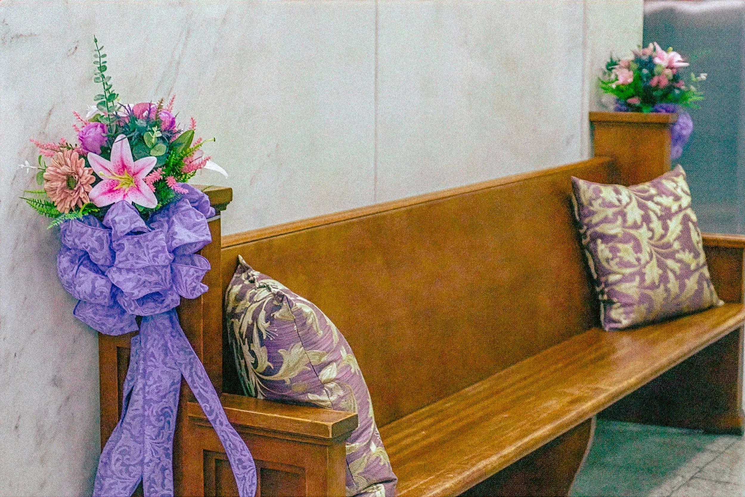 A wooden church pew with two floral arrangements placed at each end, featuring pink lilies, purple flowers, and greenery, with decorative purple ribbons, against a plain wall background.