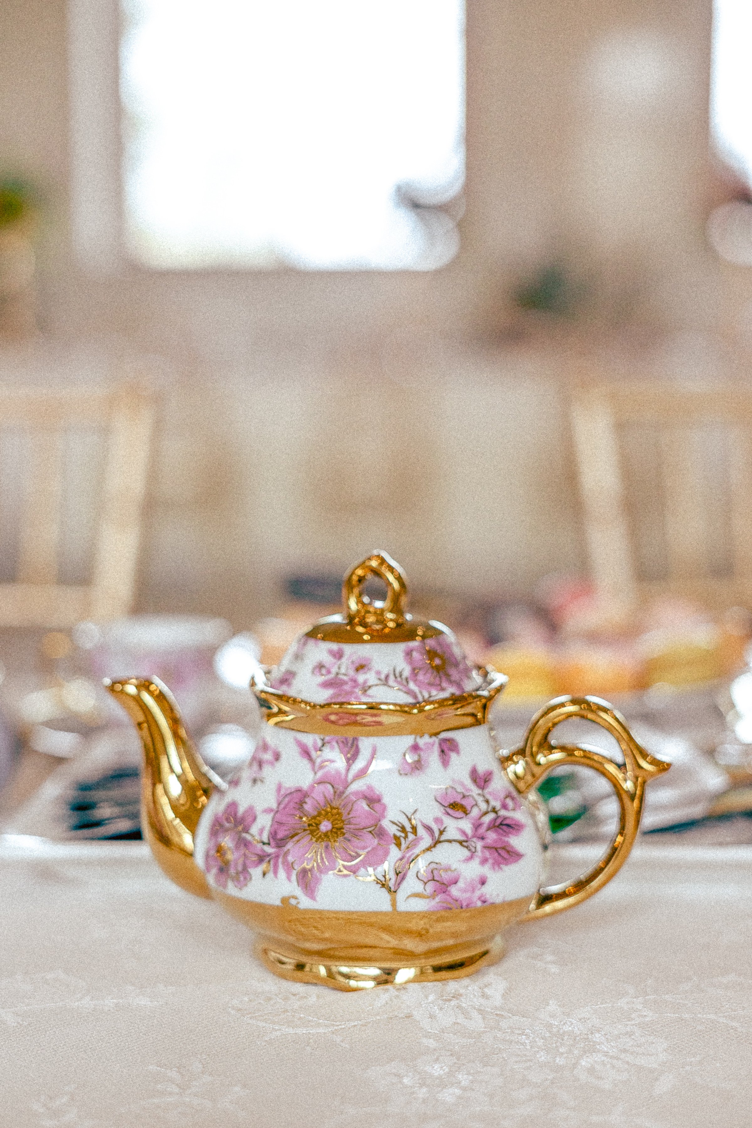 Close-up of an ornate, floral-patterned porcelain teapot with gold accents, placed on a tablecloth, with a blurry background of a room with windows.