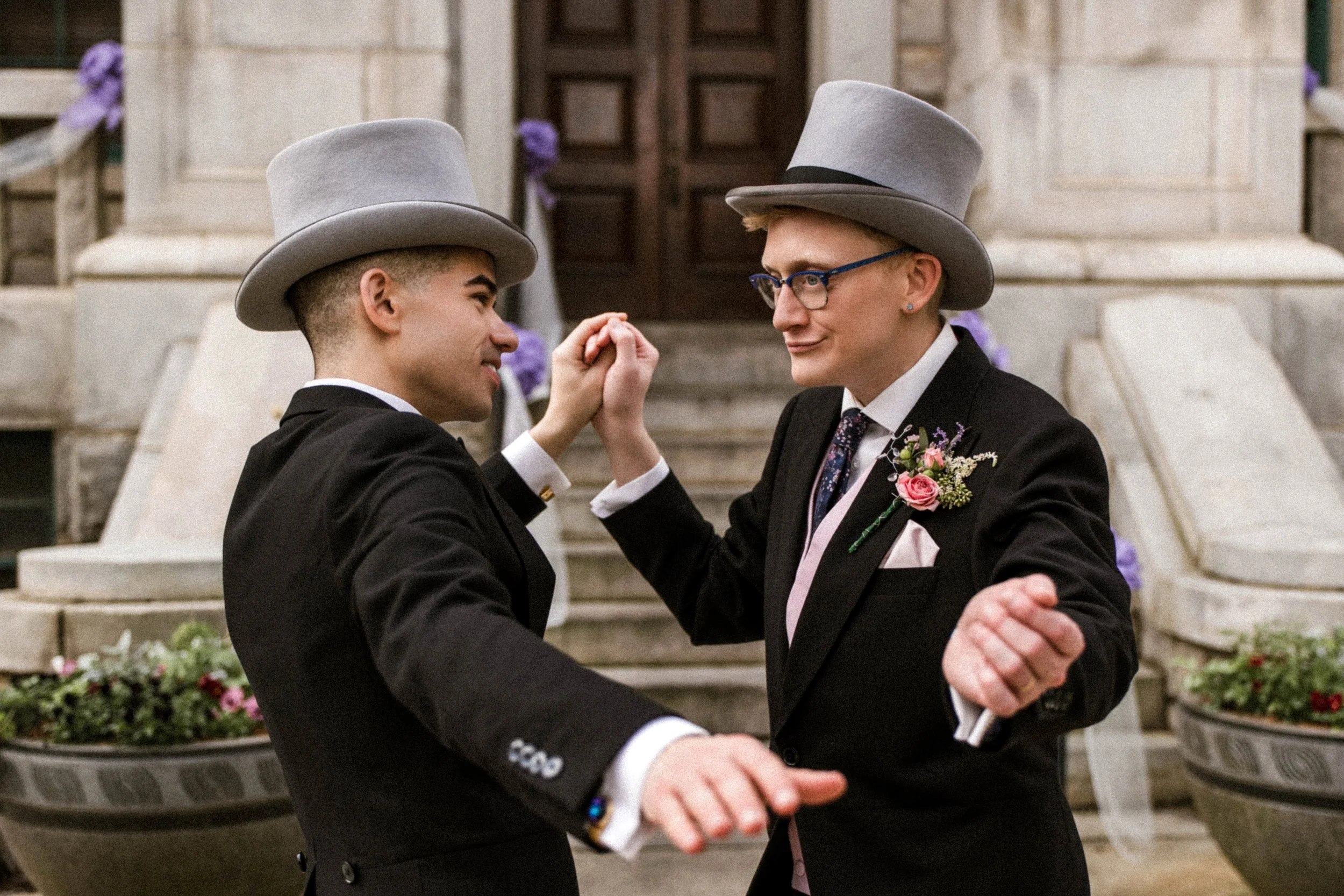 Two men in tuxedos and gray top hats dance together outside on stone steps, holding hands, with purple flowers and potted plants in the background.