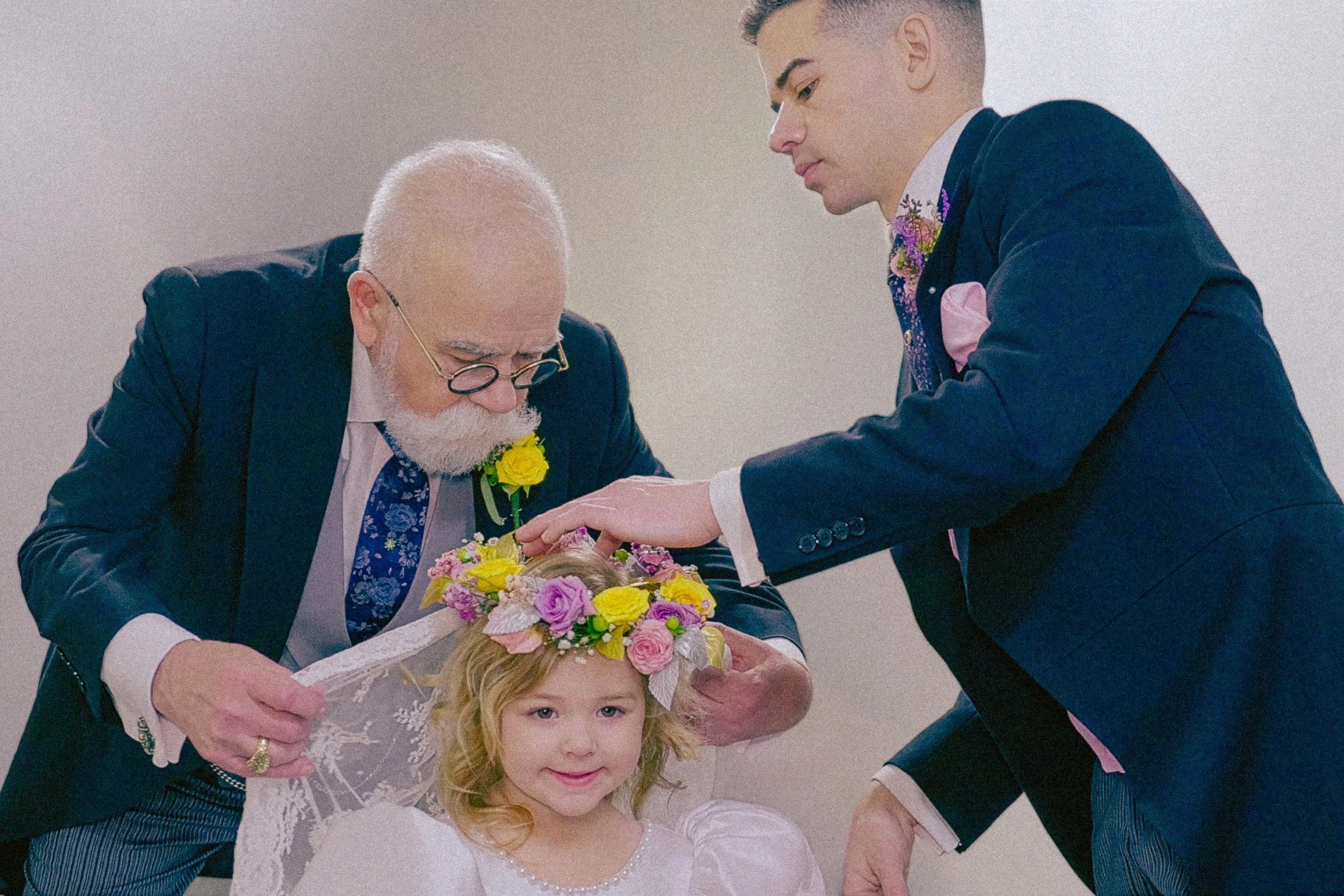 An elderly man with glasses and a white beard, wearing a dark suit and floral tie, is placing a flower crown on a young girl with blonde hair, who is dressed in a white dress. A young man in a dark suit is assisting by holding the girl's hand. The sc