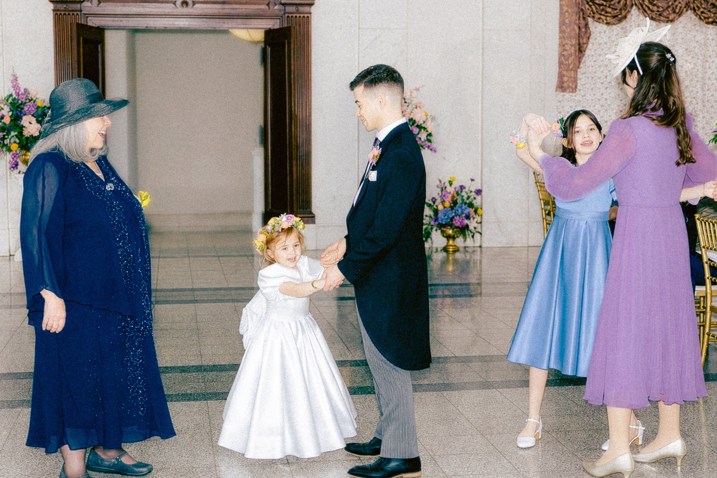 A young girl in a white dress holding hands with a man in a suit, surrounded by women dressed in vintage-style dresses and hats, at a formal event with floral decorations.