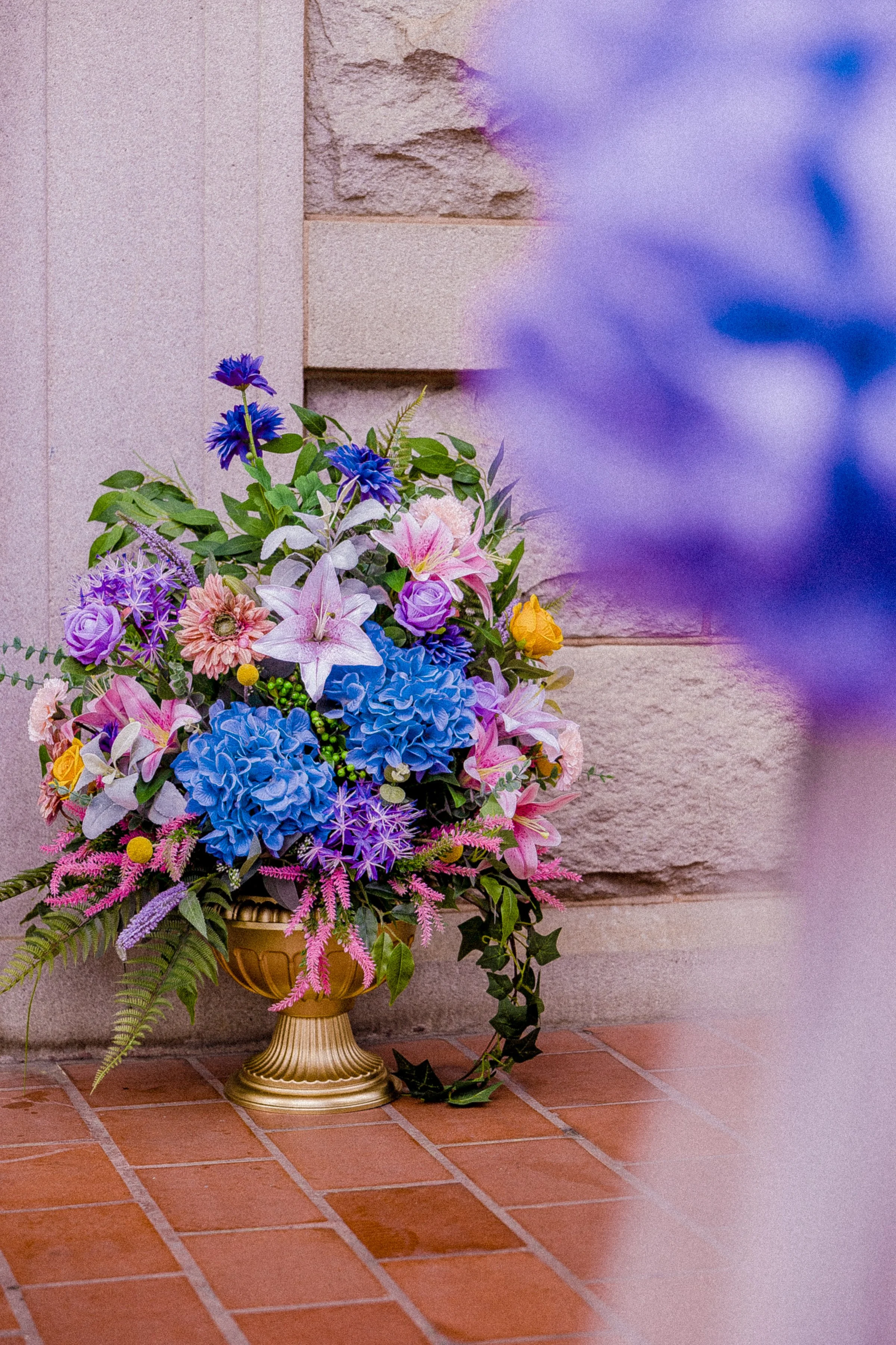 Colorful flower arrangement in a gold vase resting on a brick floor, with a stone wall in the background.