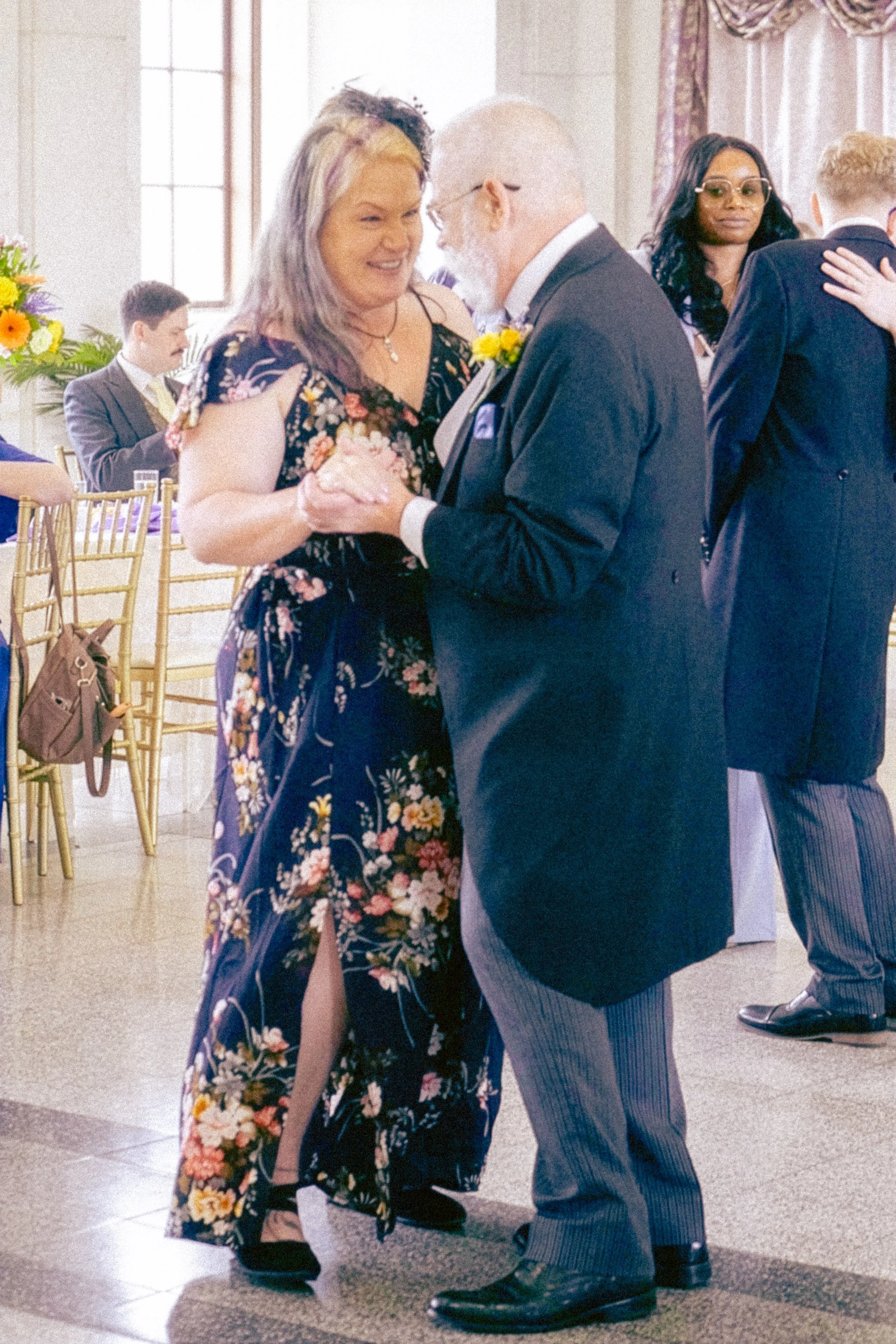 An elderly man with glasses and a white beard and a woman with shoulder-length purple hair dancing together at an indoor event, with other couples dancing in the background.