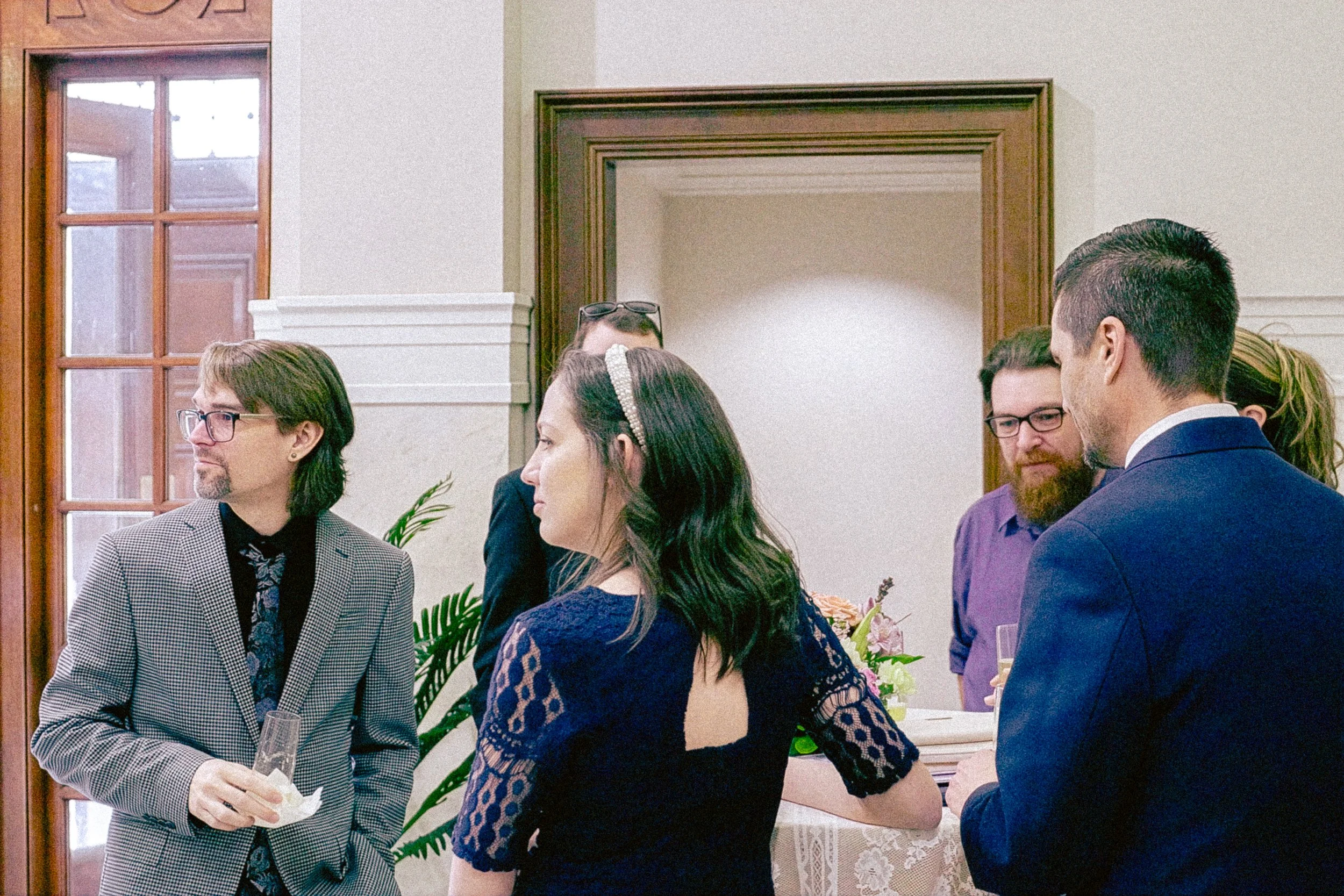 People attending a formal event or gathering in a room with wooden window frames and light-colored walls. Some individuals are holding glasses, and there are floral arrangements in the background.