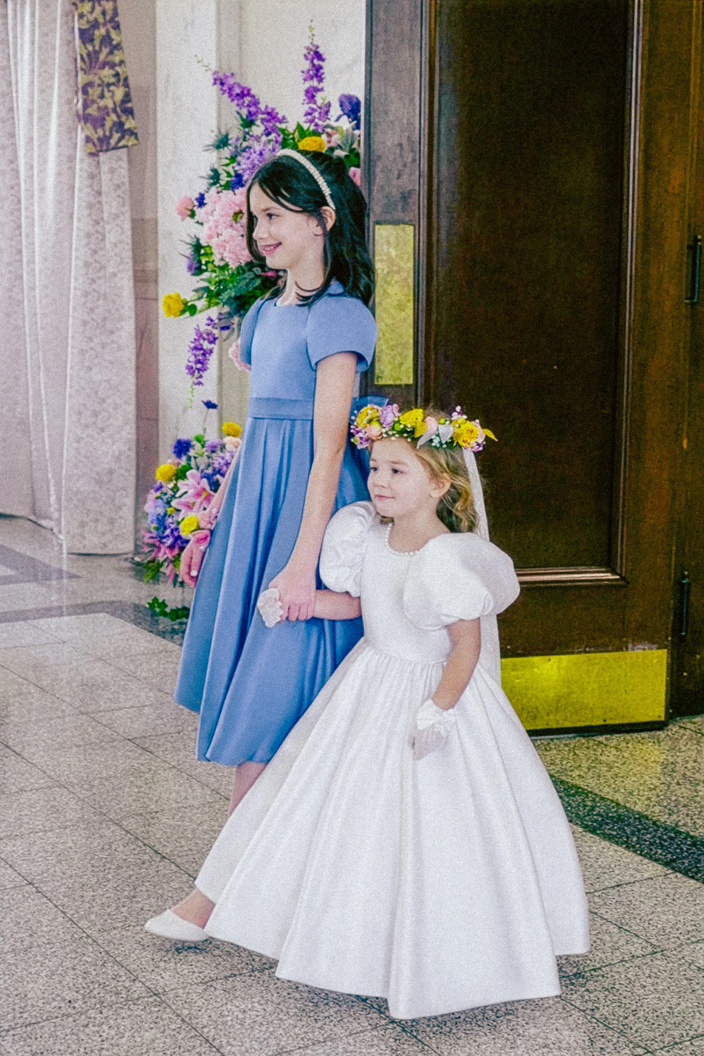 A young girl in a white dress with a floral crown holding hands with a woman in a blue dress with a pearl headband, standing in a decorated indoor space with flowers and patterned curtains.
