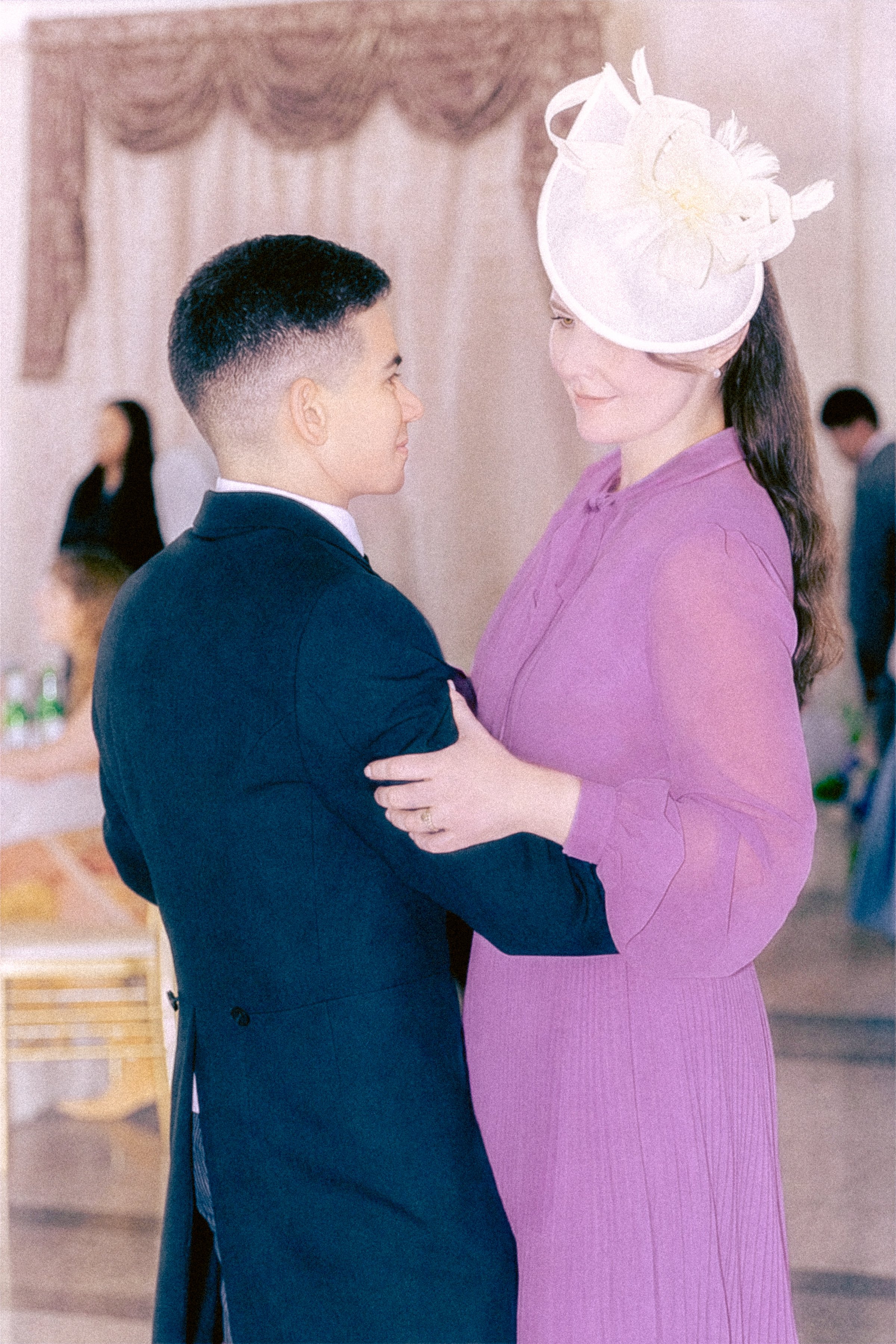 A man and a woman are dancing closely together at a social event. The man is wearing a dark suit and the woman is wearing a purple dress with a decorative hat. They are facing each other and maintaining eye contact.