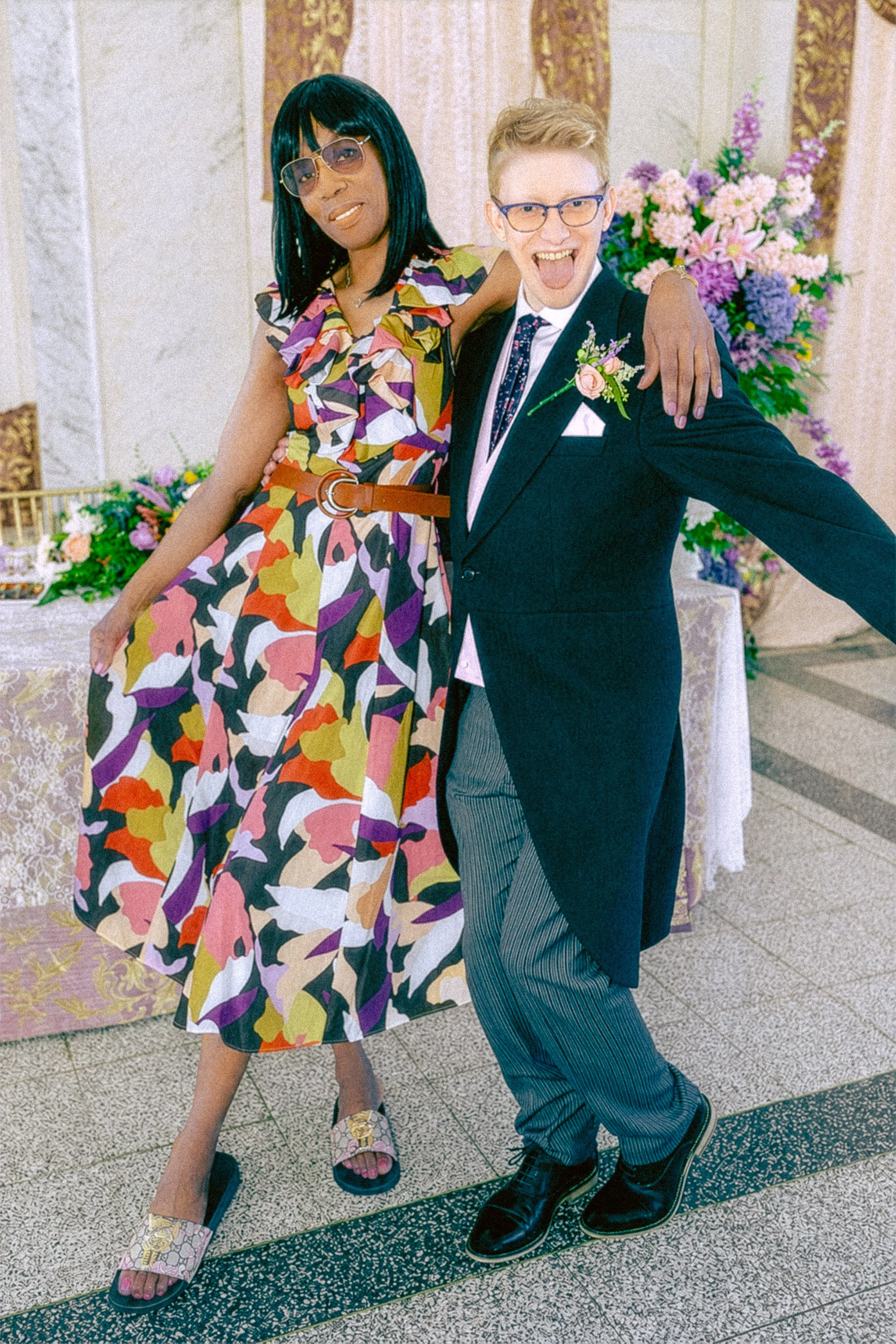 A woman in a colorful patterned dress with glasses and sandals and a man in a dark tuxedo with striped pants, glasses, and dress shoes. They are standing close together, smiling and celebrating at a decorated event with pink and purple flowers in the