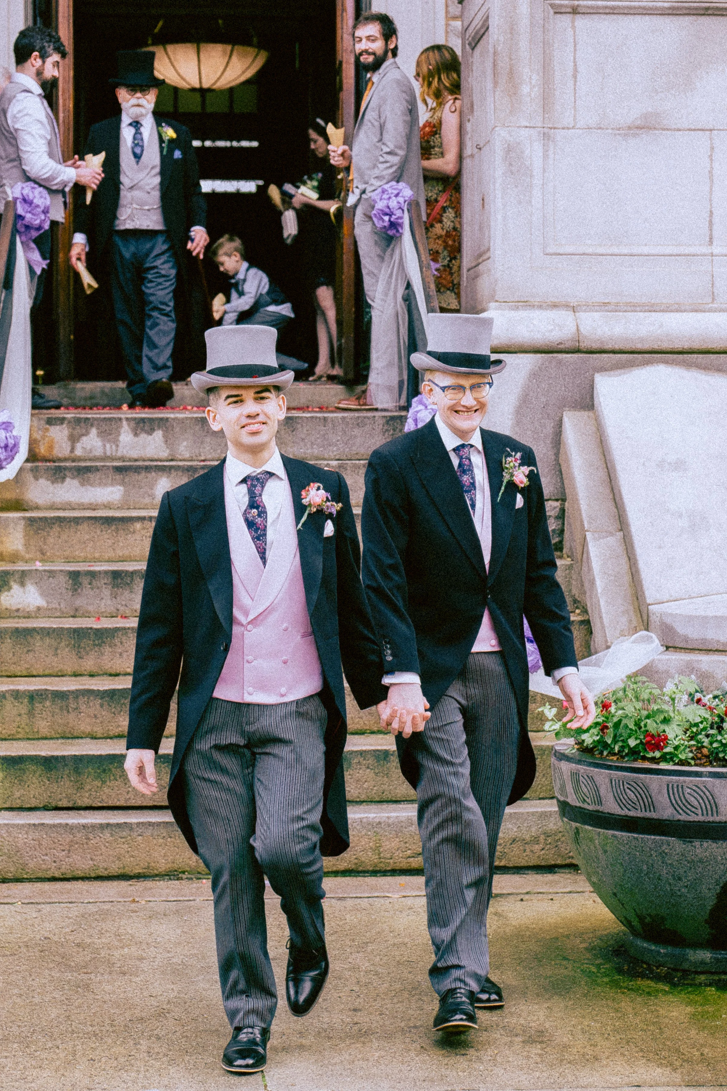 Two men dressed in traditional black tailcoats, pink vests, and striped trousers, holding hands and walking down steps. They are wearing top hats and boutonnières. In the background, a wedding party stands outside a building, with some individuals in
