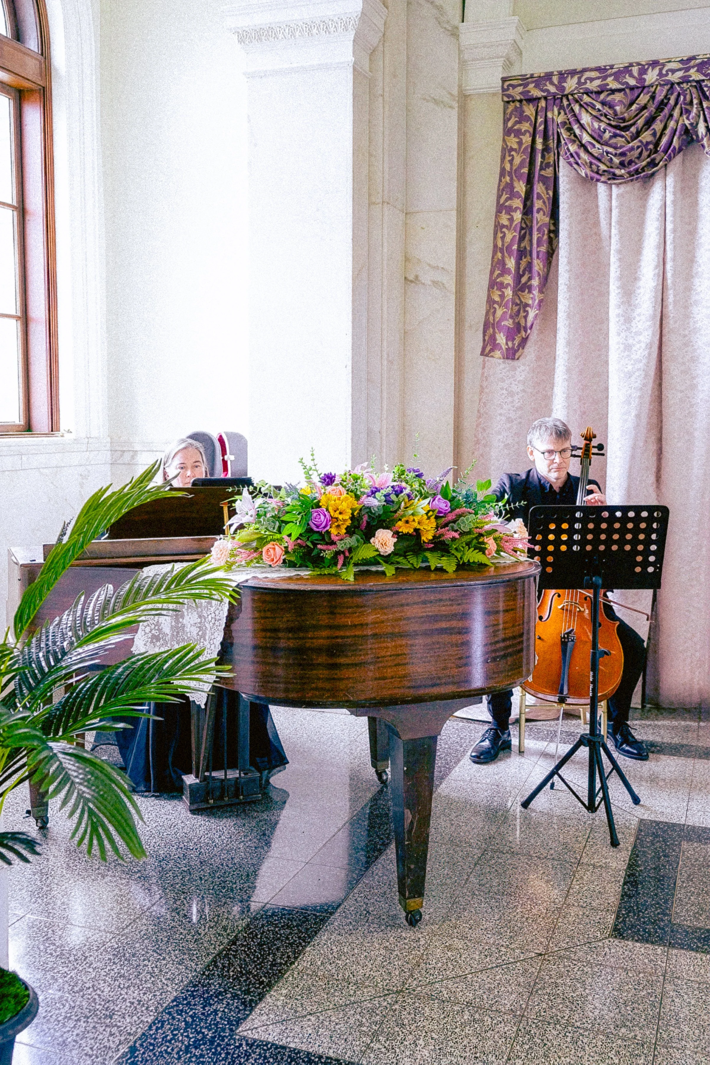An elegant room with high ceilings, large window, and ornate curtains. In the foreground, a grand piano with a colorful flower arrangement on top. Behind the piano, a woman is partially visible, and a man with glasses is playing a cello. A music stan