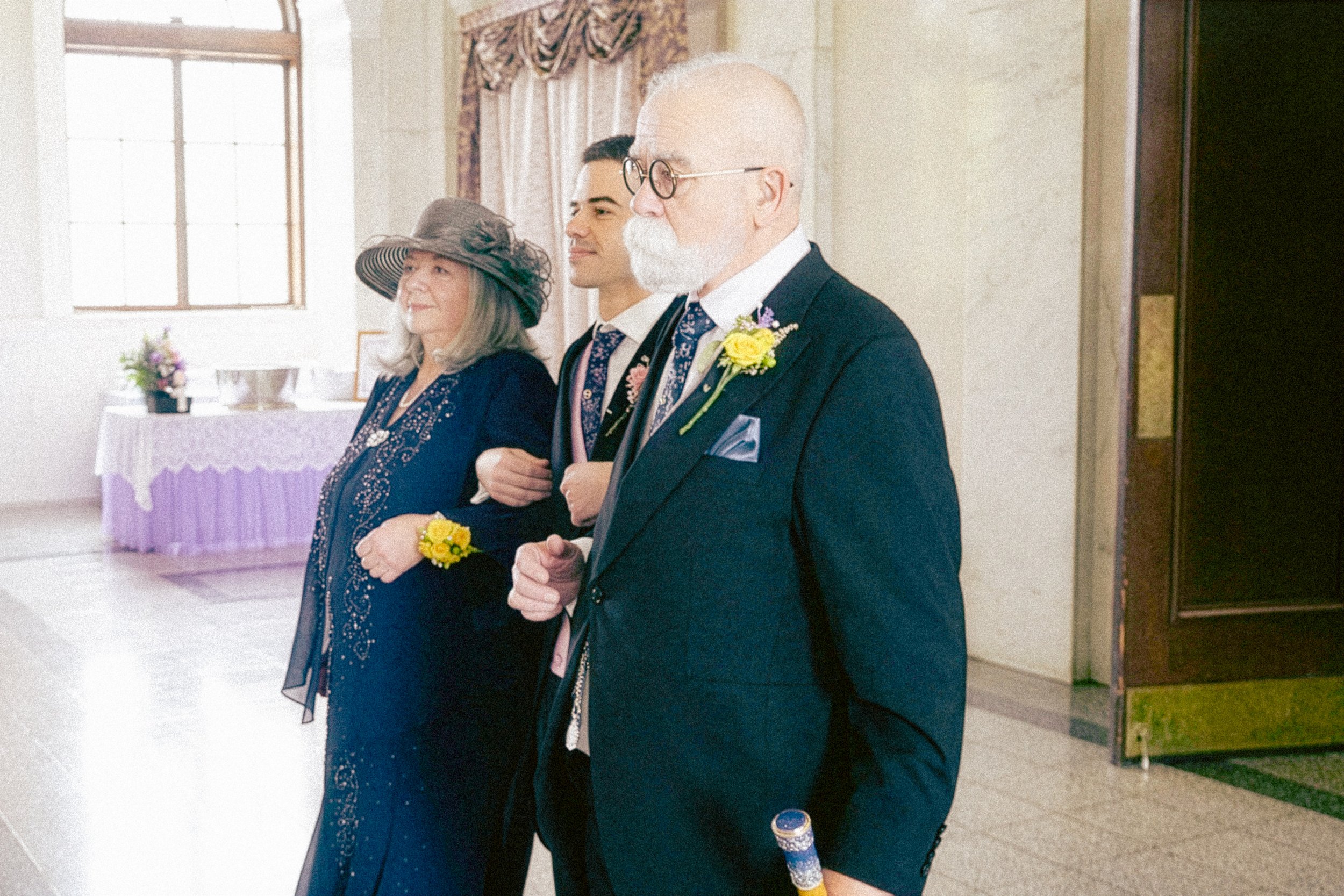 Three people standing side by side inside a room, dressed in formal attire, with candles and a purple tablecloth in the background.