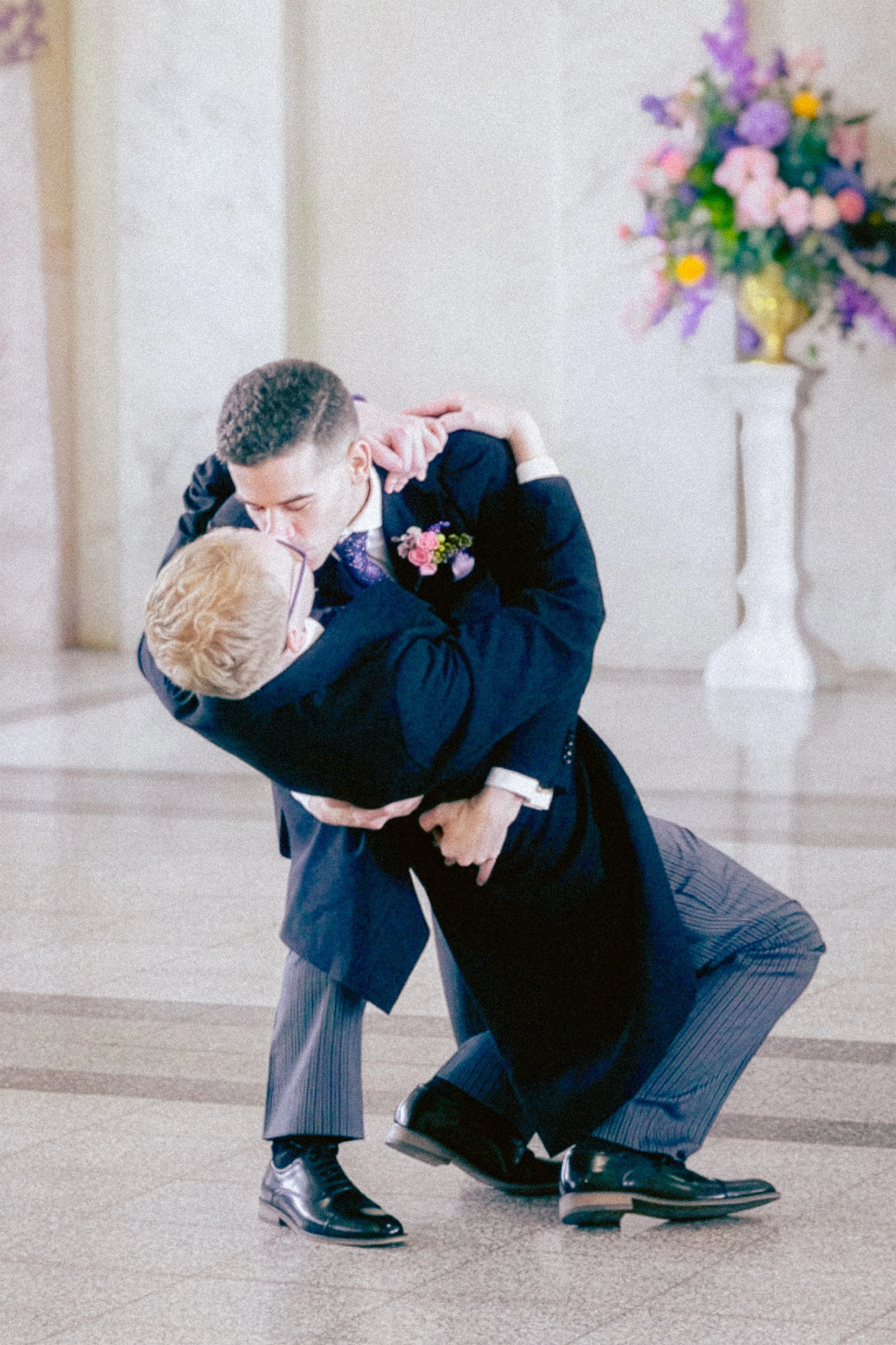 Two men in formal attire dancing passionately indoors, with a large colorful bouquet of flowers in a white vase in the background.