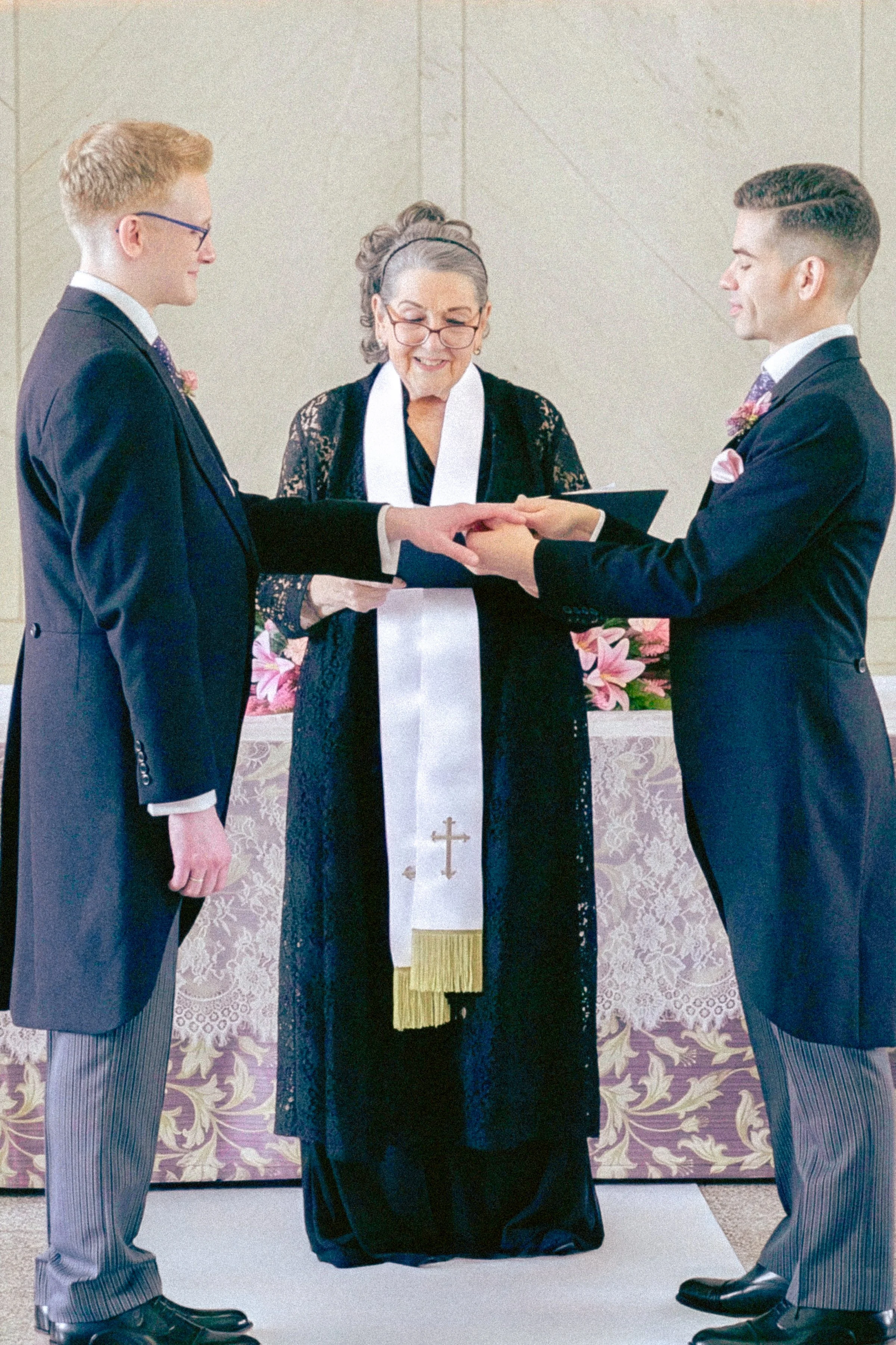 Two men dressed in suits holding hands during a wedding ceremony with a woman officiating.