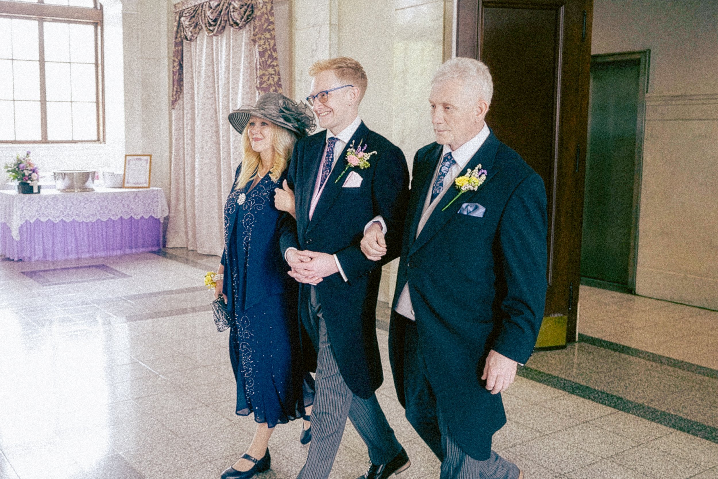 Three people walking arm-in-arm in an indoor setting, dressed in formal attire, with floral corsages, indicating they are attending a special event such as a wedding or celebration.