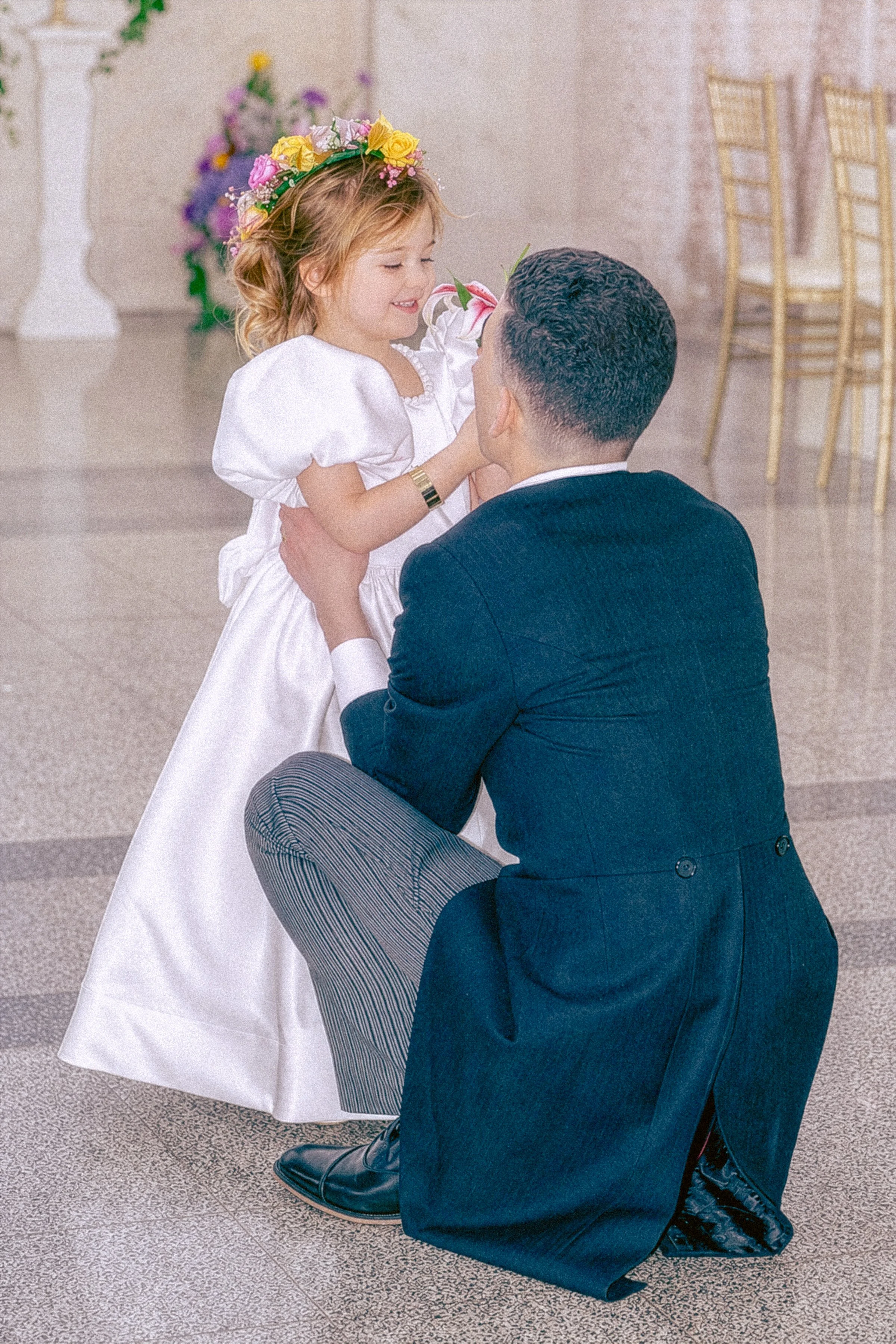 A man kneeling on one knee holding a young girl in a white dress with a floral crown, inside a decorated hall with chairs and floral arrangements.
