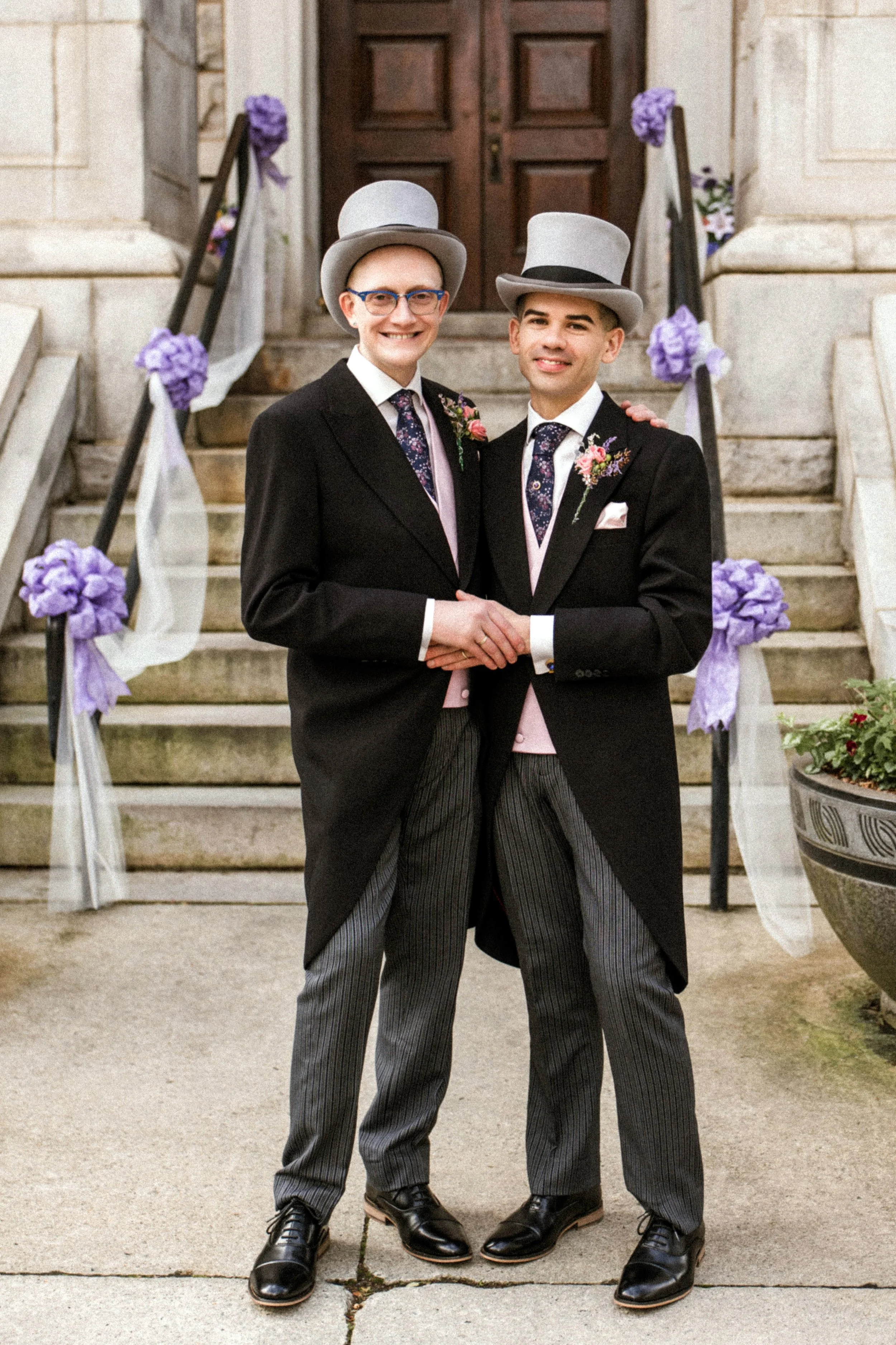 Two men in tuxedos and top hats standing on outdoor stone steps decorated with purple flowers and tulle, smiling and holding hands at a formal event.