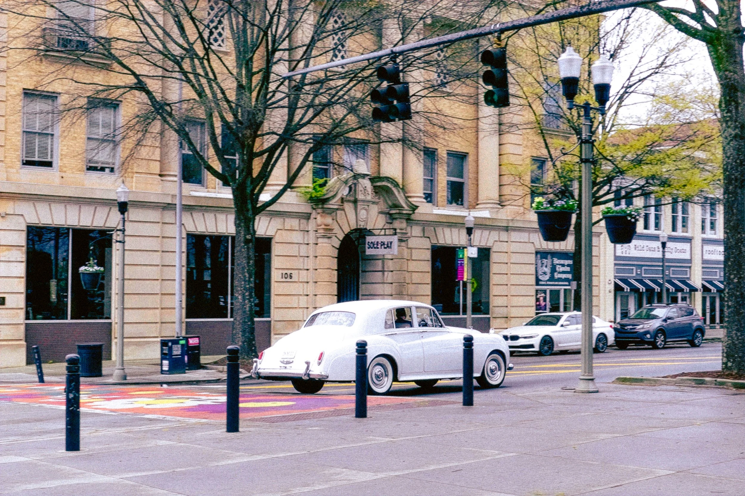 Street scene with vintage white car, modern vehicles, trees, streetlights, and buildings with large windows and historical architecture.
